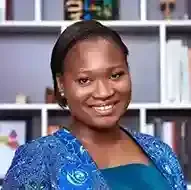 A woman in a blue jacket is smiling in front of a bookshelf.