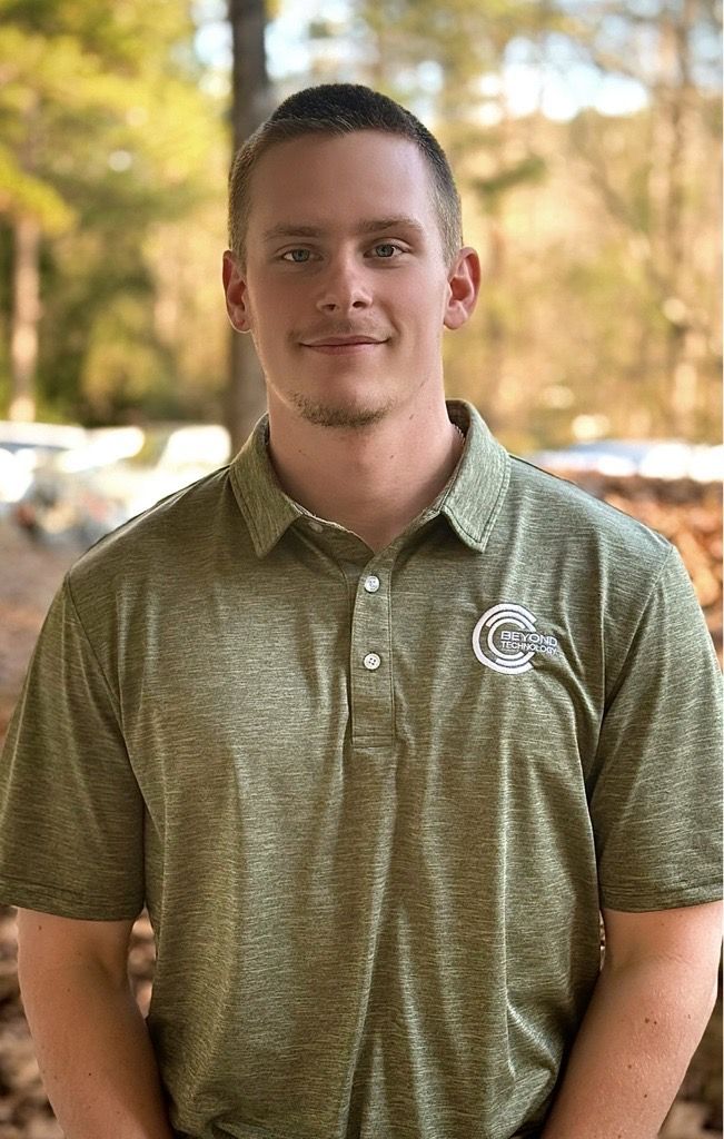 Braedon Graham, IT Technician at Beyond Technology, wearing a textured olive-green polo shirt stands outside in a wooded setting, looking forward with a neutral smile.