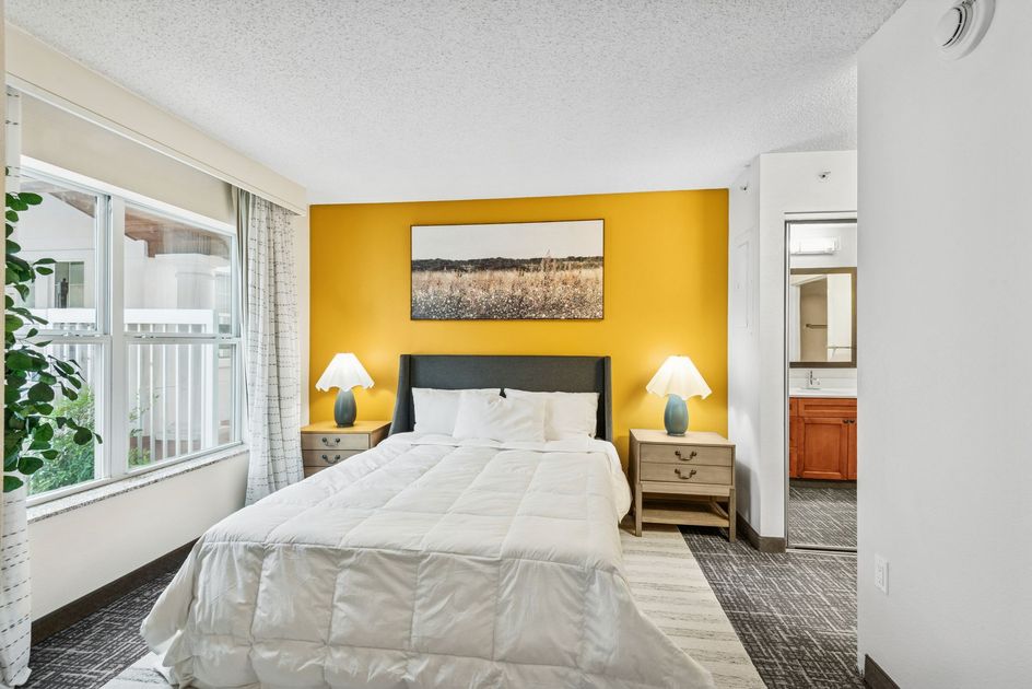 Bedroom with yellow accent wall, bed, nightstands, window, and doorway to a bathroom.
