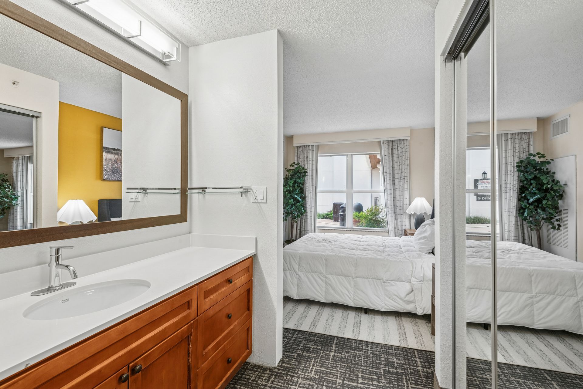 Bathroom with vanity and mirror, looking into bedroom with bed and window.