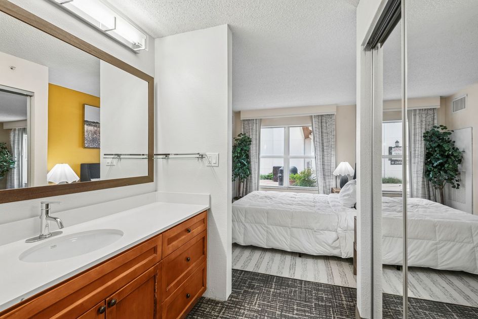 Bathroom with vanity and mirror, looking into bedroom with bed and window.