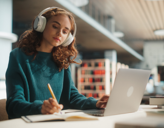 Woman in headphones writing in a notebook, using a laptop in a library.