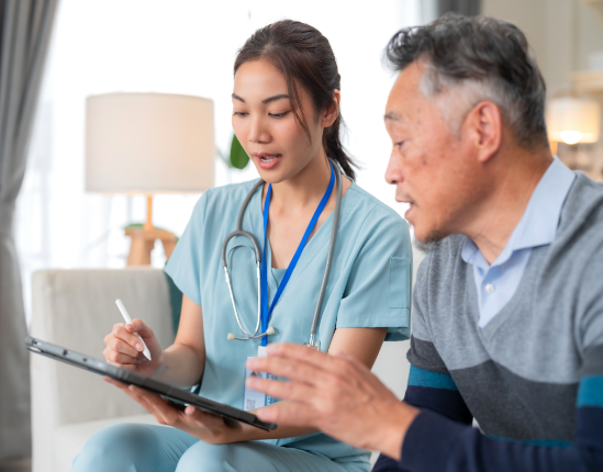 Nurse in blue scrubs points at a tablet while talking to an older Asian man in a living room.