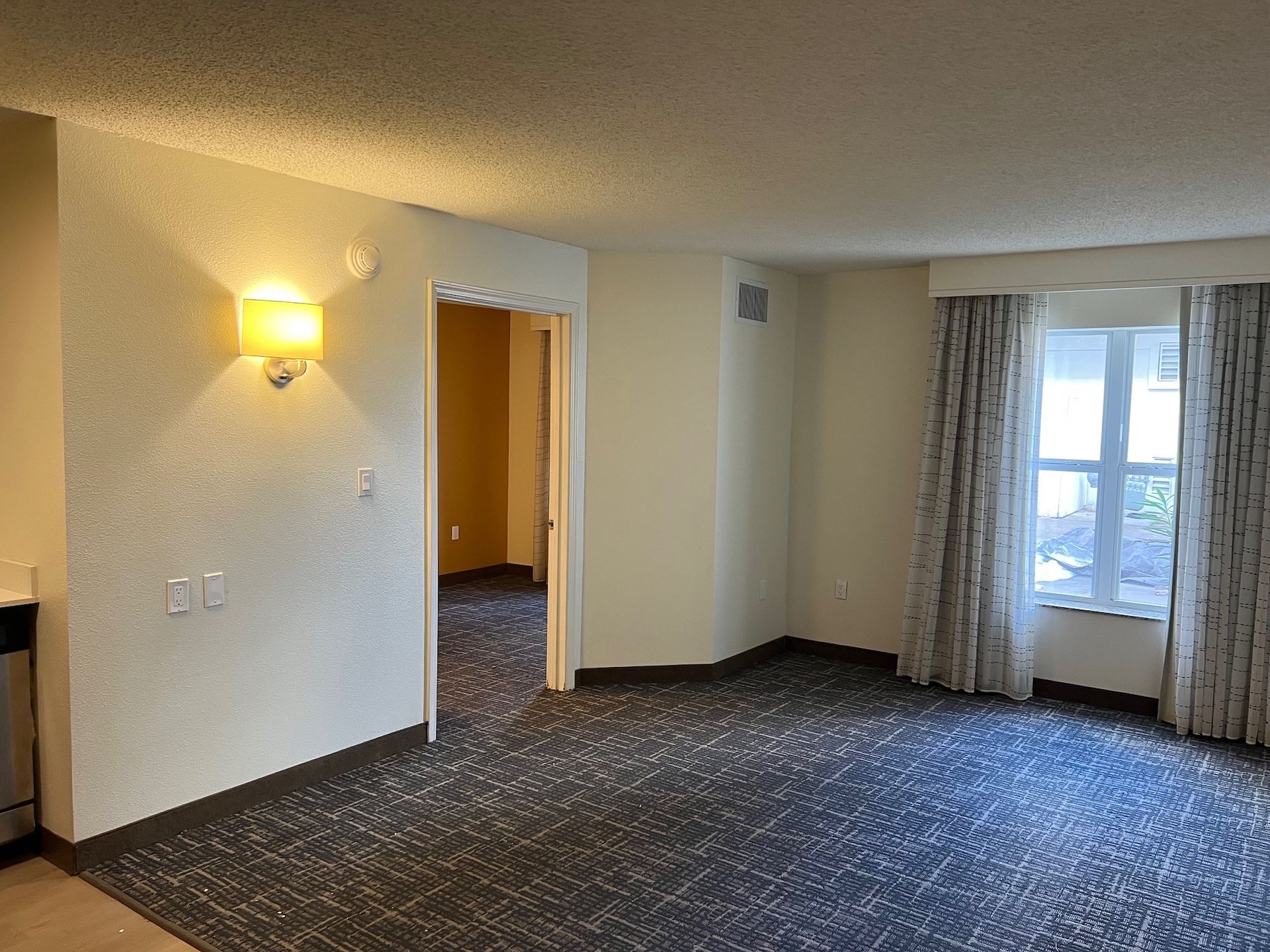 Empty living room with patterned carpet, doorway to another room, window with curtains, and wall sconce.