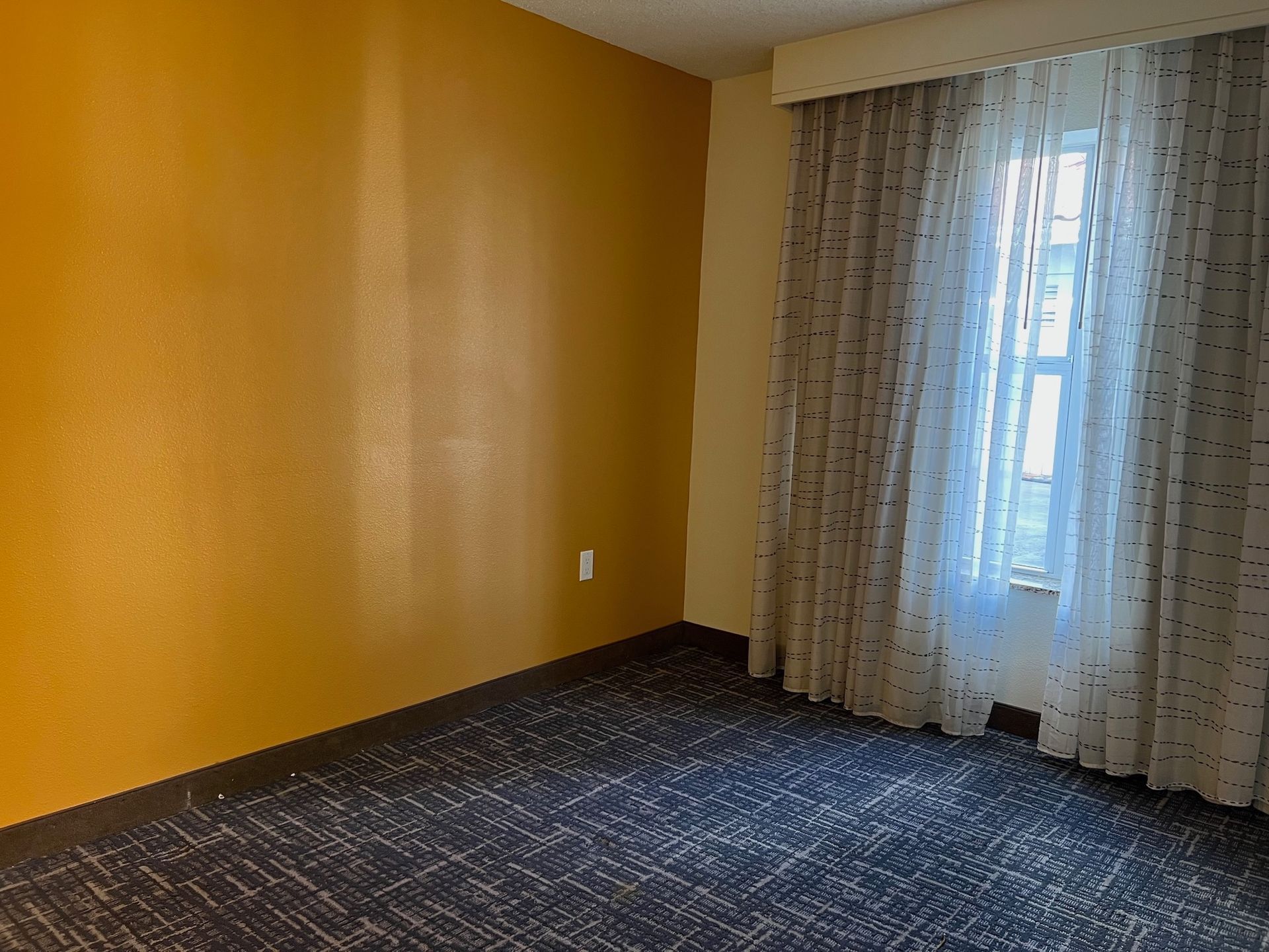 Empty room with mustard-colored wall, patterned carpet, and sheer curtains over a window.