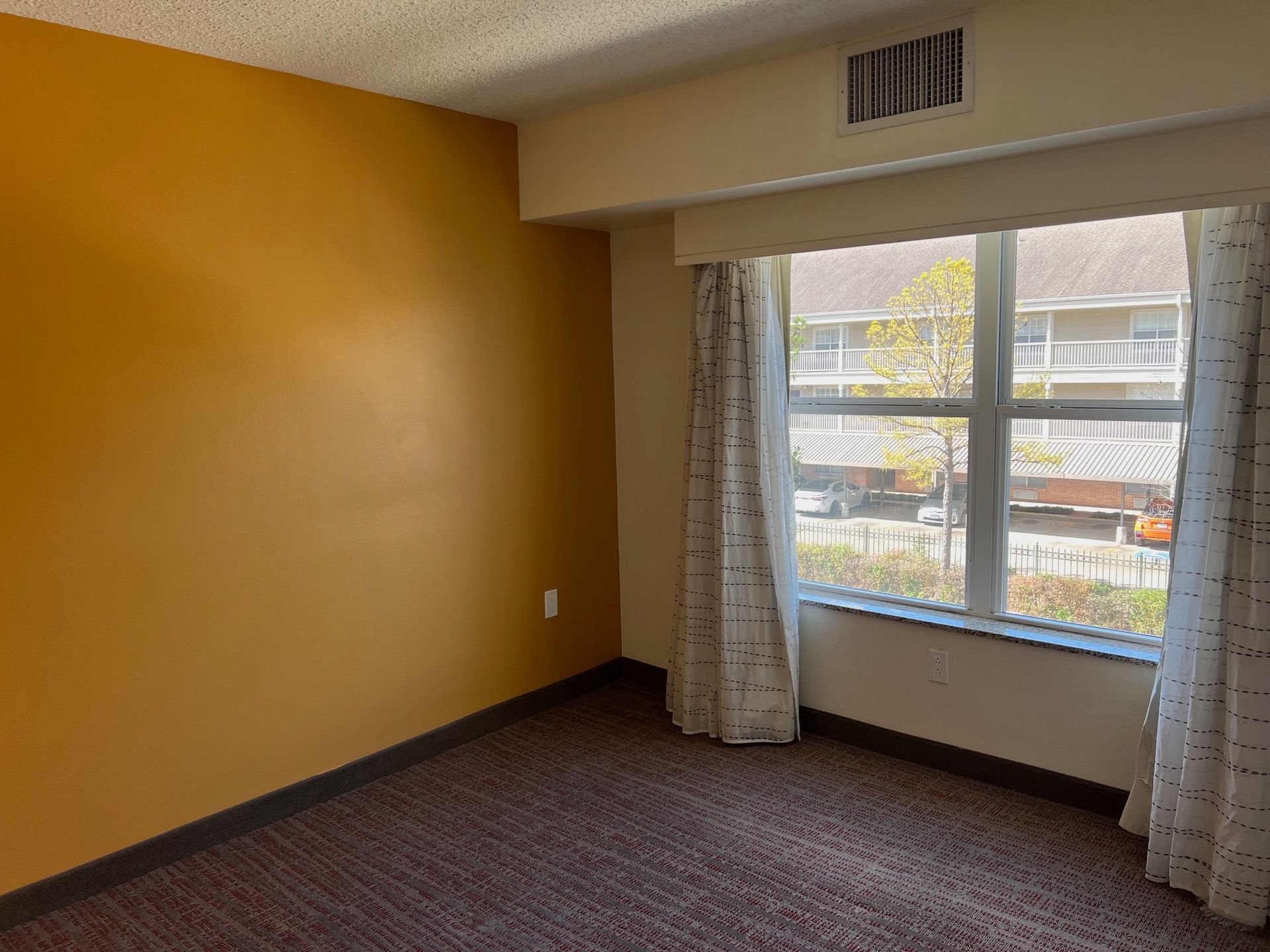 Empty room with gold wall, window with sheer curtains, and brown carpet.