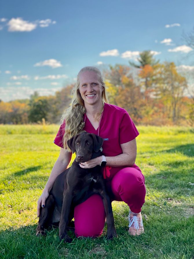 Dr. Hoffman pink scrubs kneeling in a grassy field, smiling while holding a chocolate Labrador retriever.