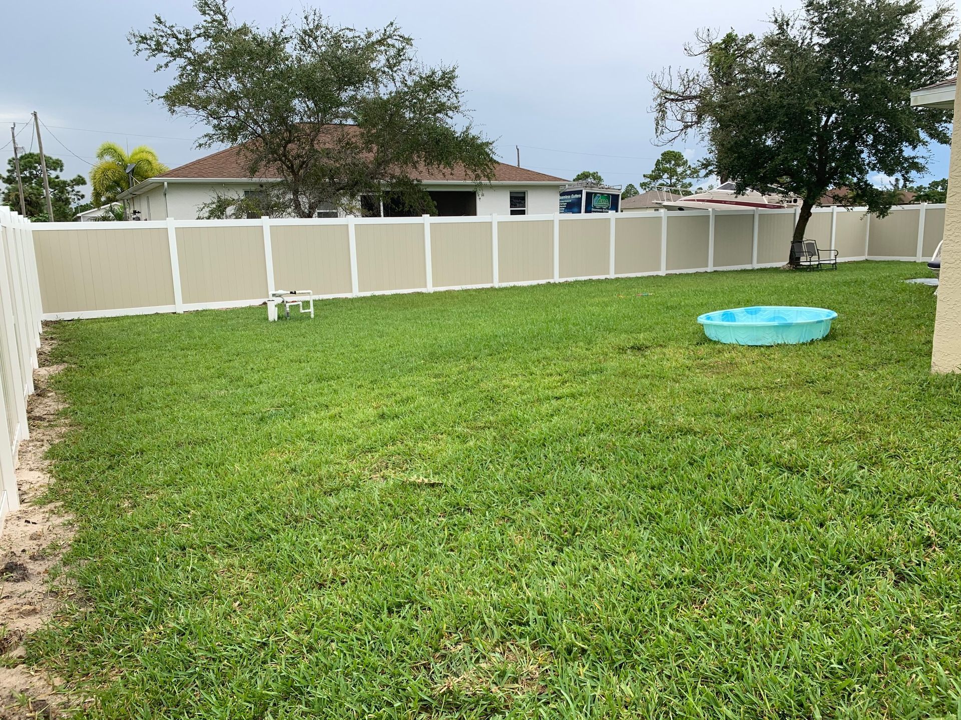 A grassy backyard with a tan and white fence, a small pool, and a tree.