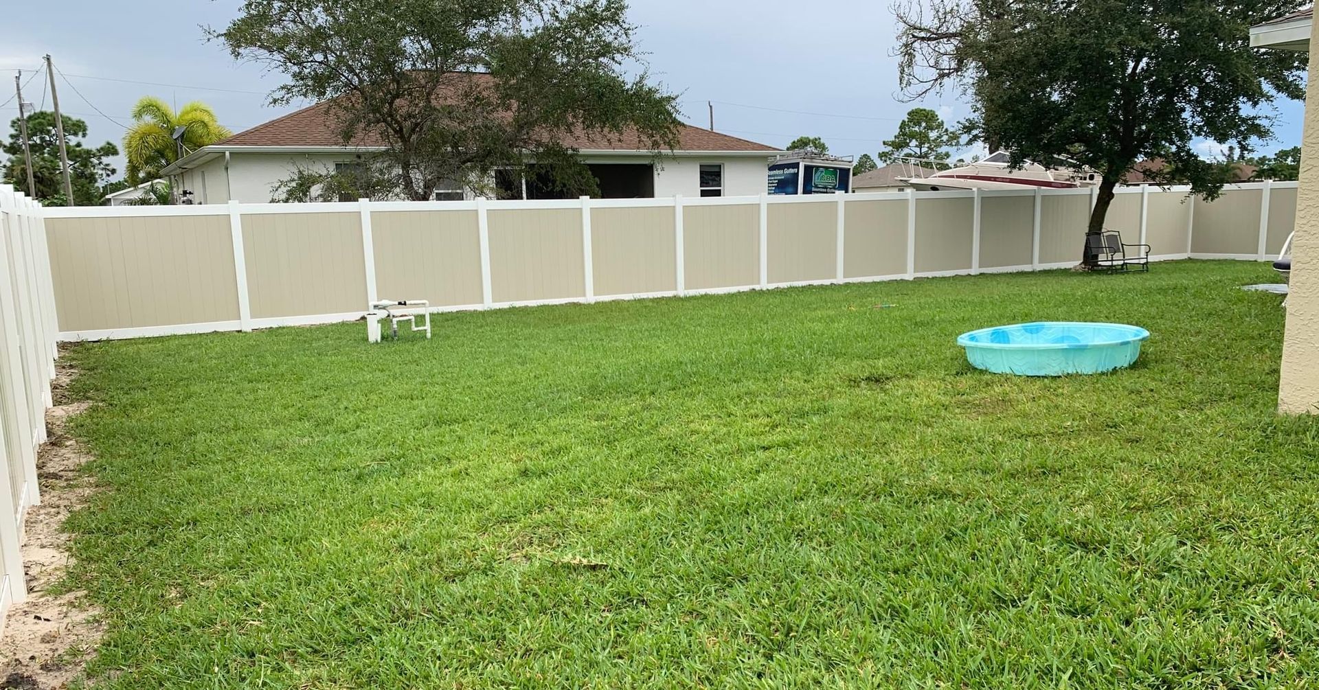 Green backyard with beige fence and small pool, a tree, and a house in the background.