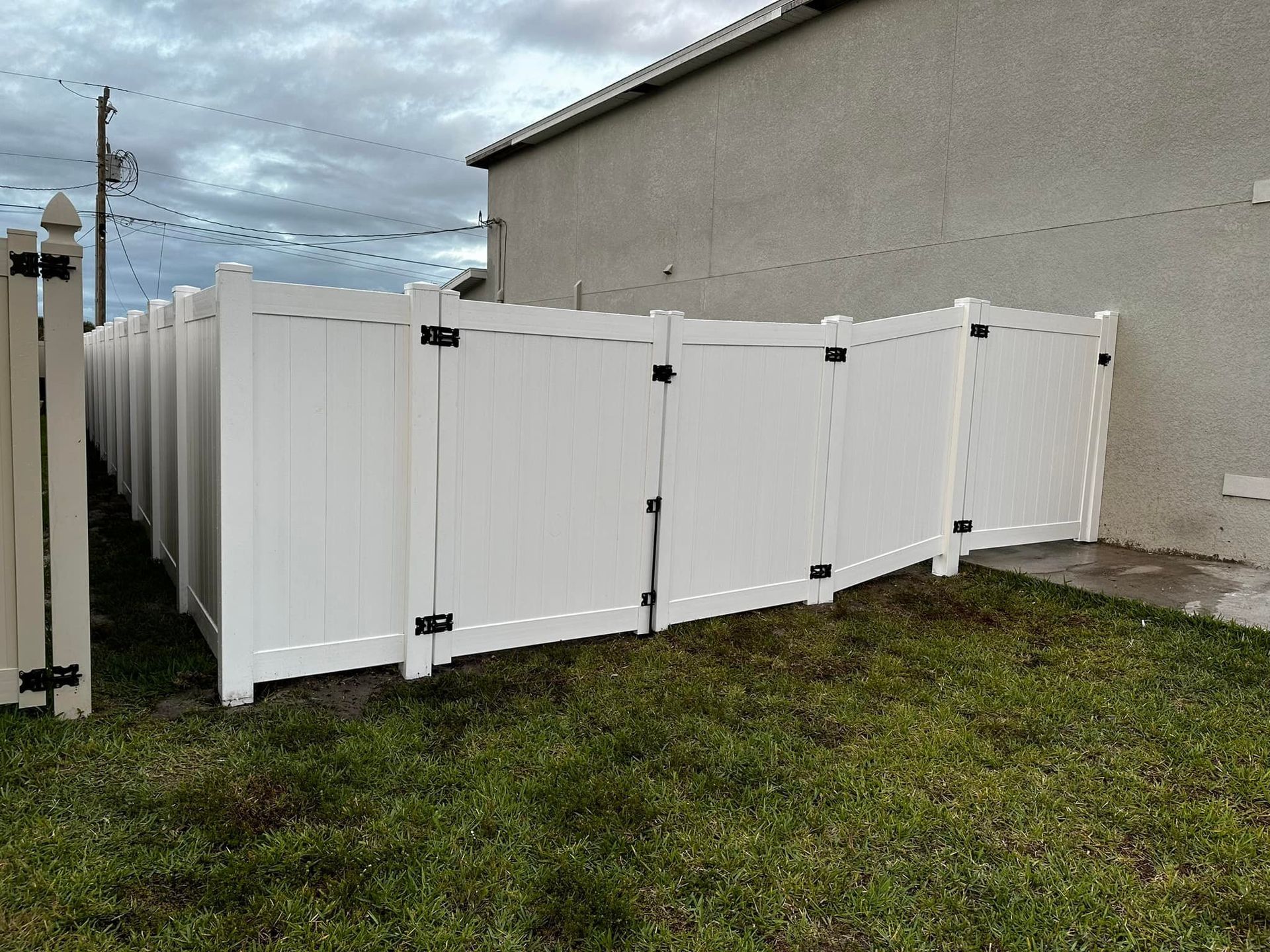 White vinyl fence against a tan building. Black hardware. Green grass. Overcast sky.