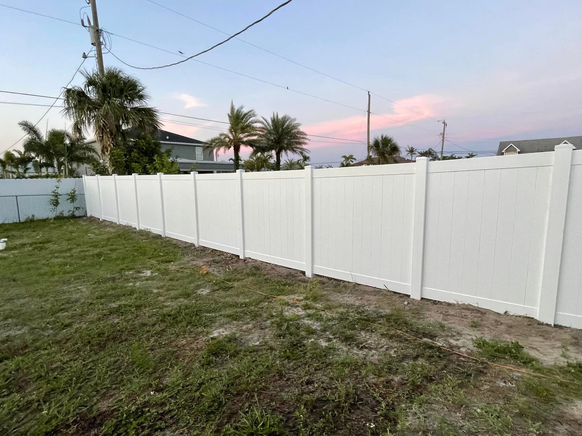 White vinyl fence encloses a grassy yard against a backdrop of trees and a pink-tinged sky.