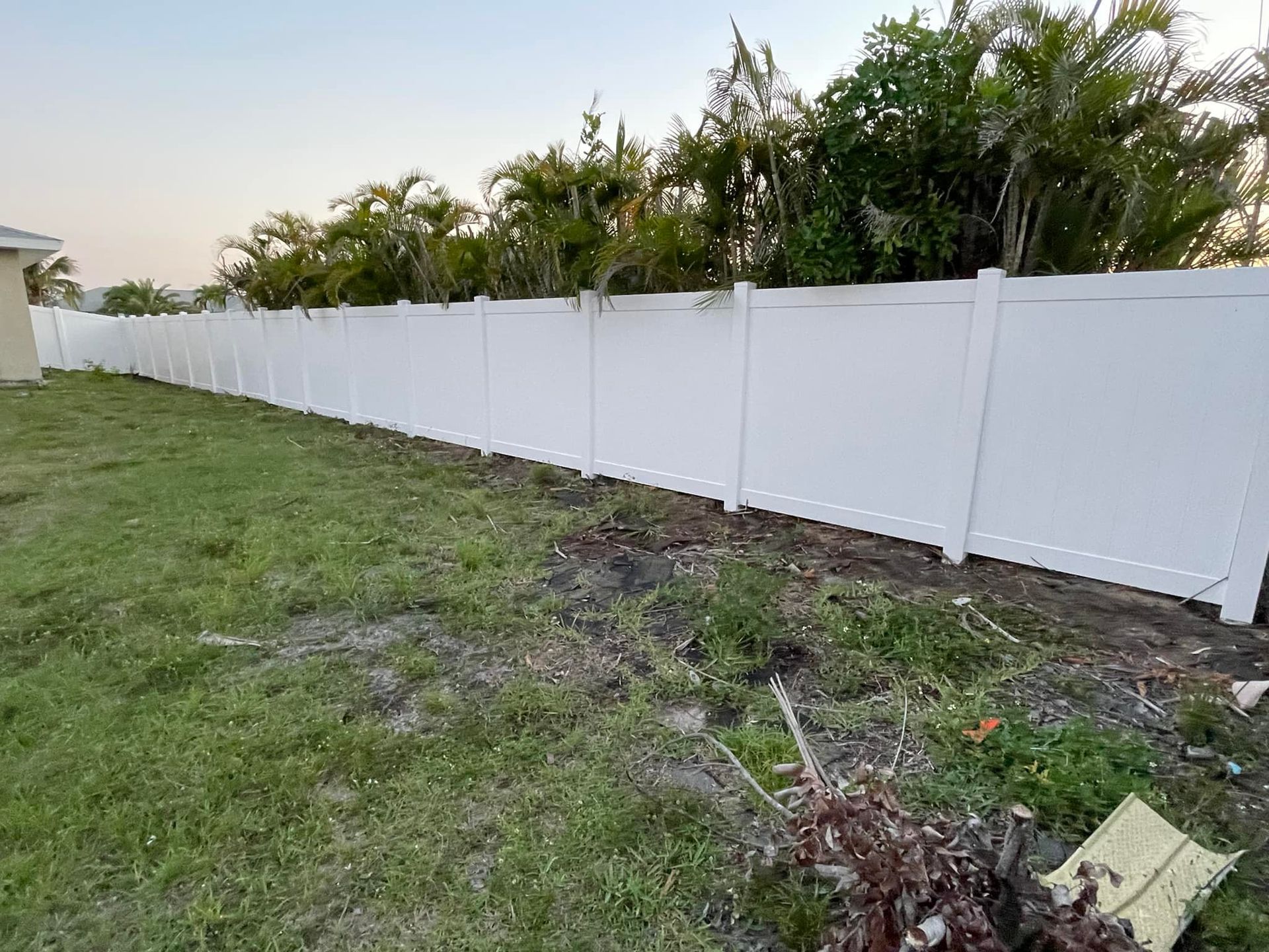 White vinyl fence bordering a grassy yard with overgrown grass, with trees in the background.