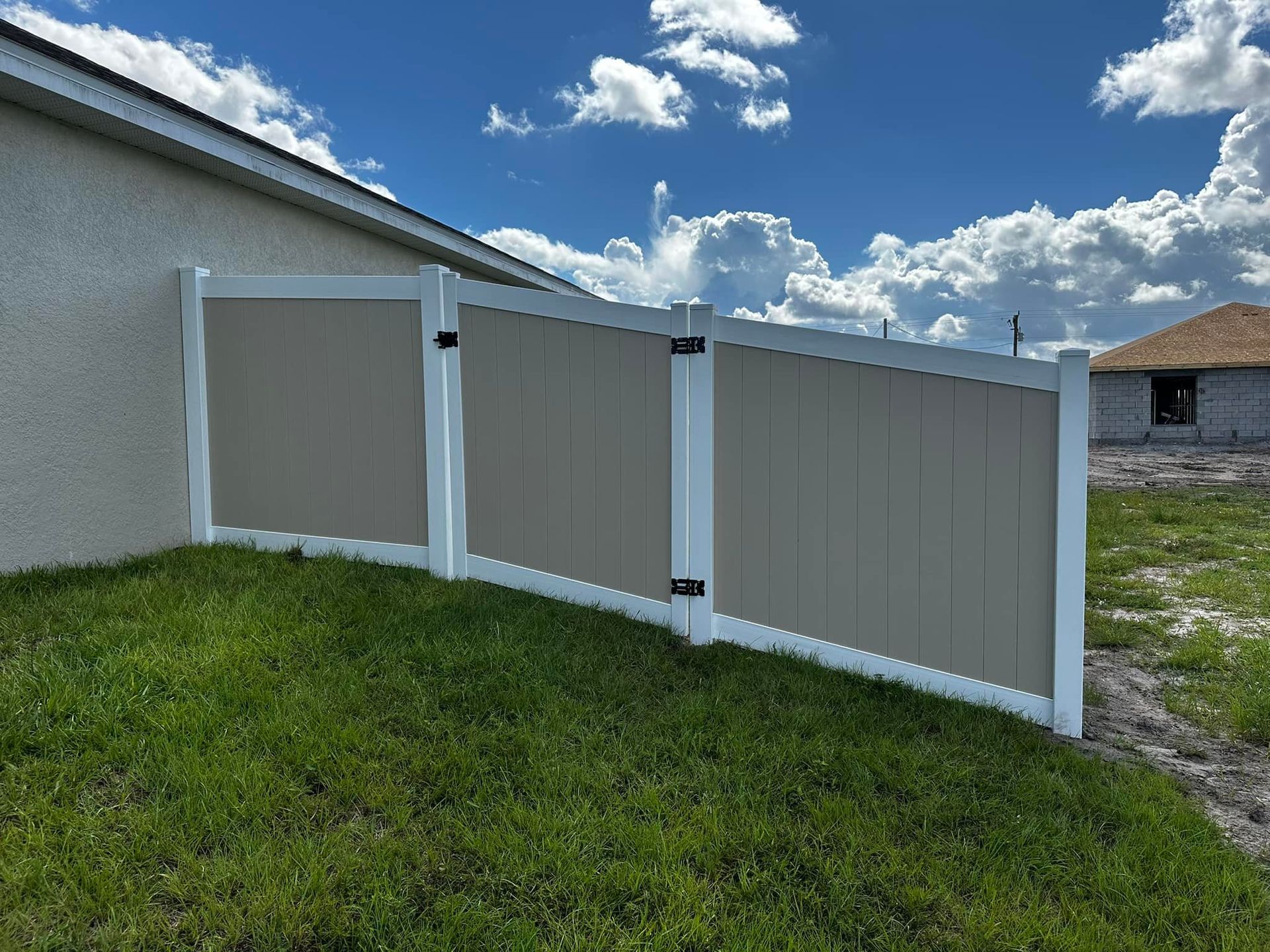 Tan and white vinyl fence along a grassy slope near a house under a blue sky.