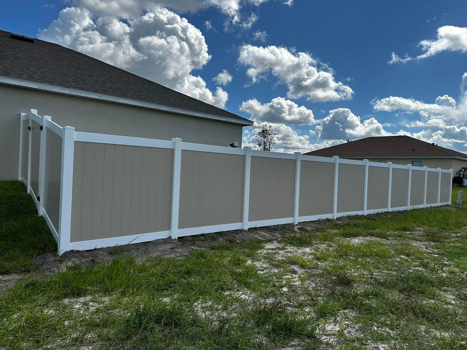 Tan and white vinyl fence along the side of two houses, under a blue sky with fluffy clouds.