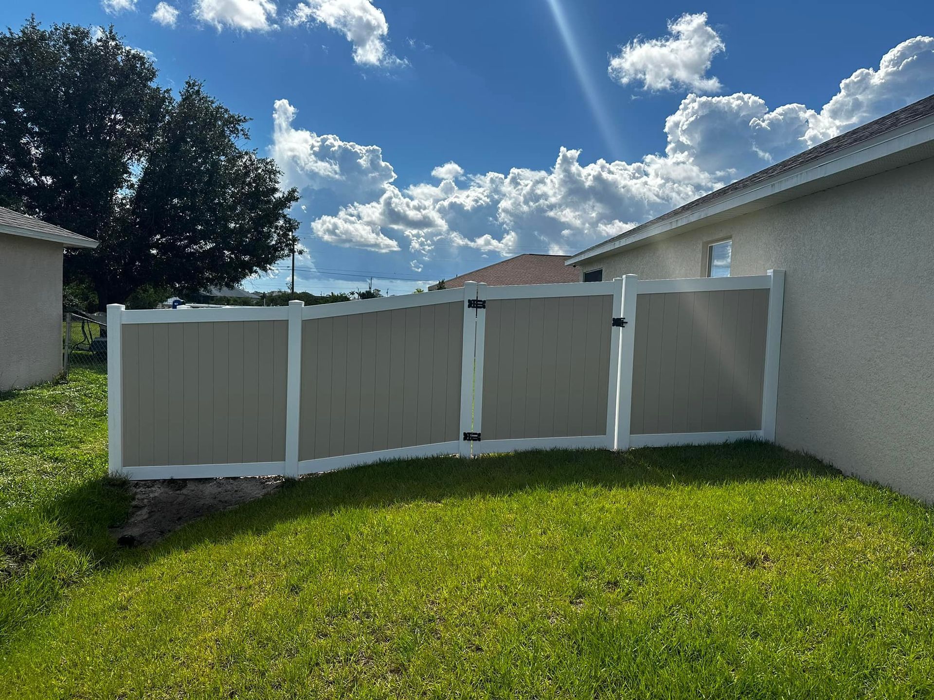 Tan and white vinyl fence in a grassy backyard on a sunny day with puffy clouds.