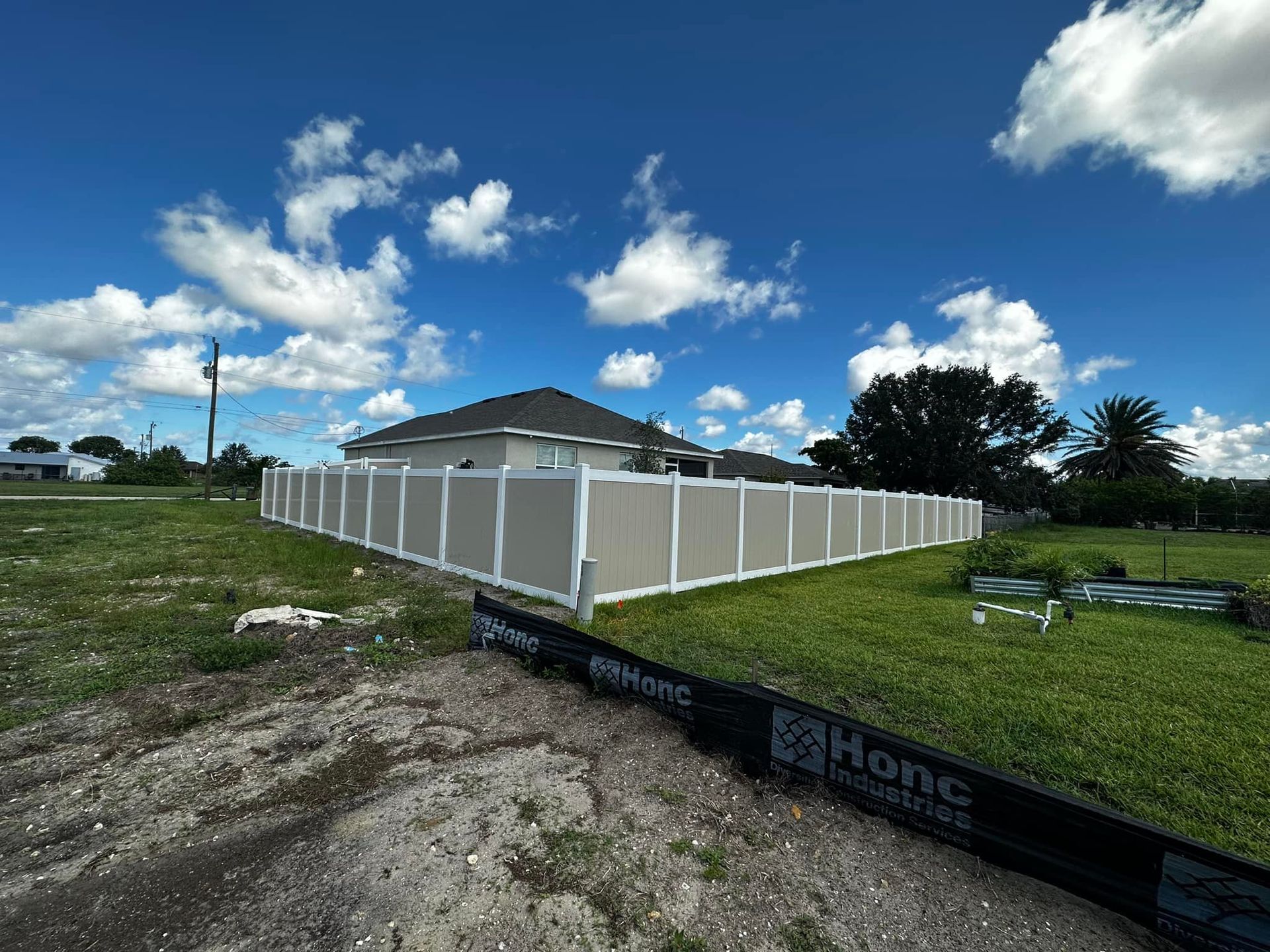 Tan vinyl fence surrounding a light-colored house on a grassy lot under a blue sky with clouds.