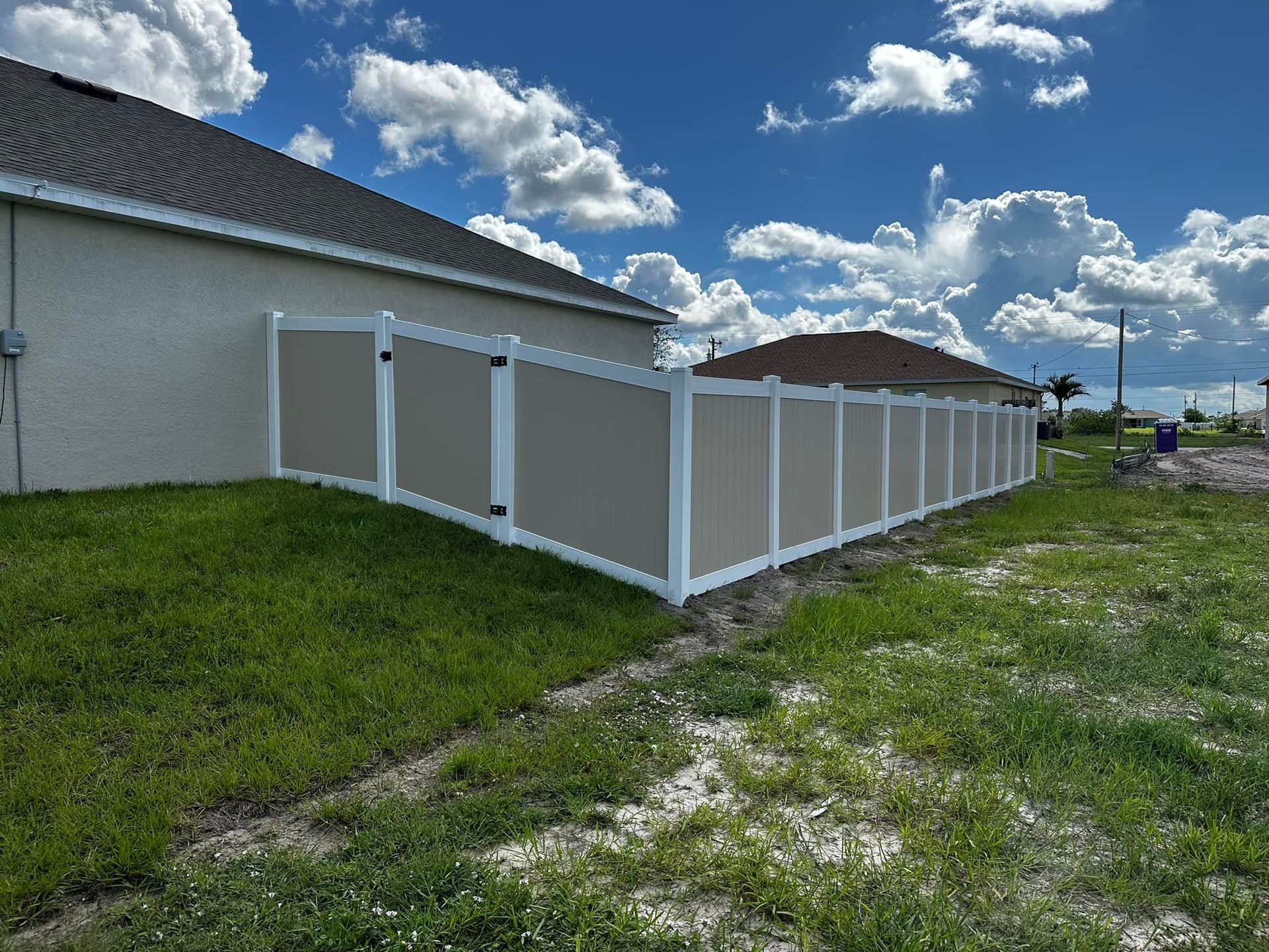 Tan and white vinyl privacy fence surrounding a house on a grassy lot under a blue sky with clouds.