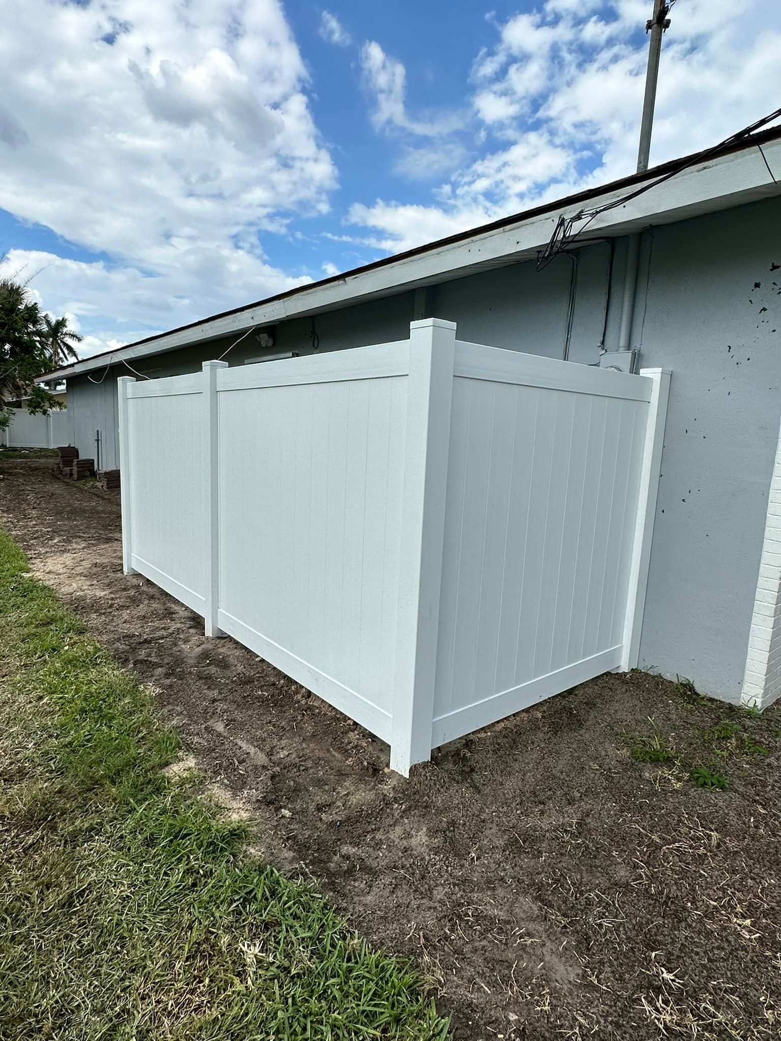 White vinyl fence against a pale building, set in a patch of dirt and grass under a cloudy sky.