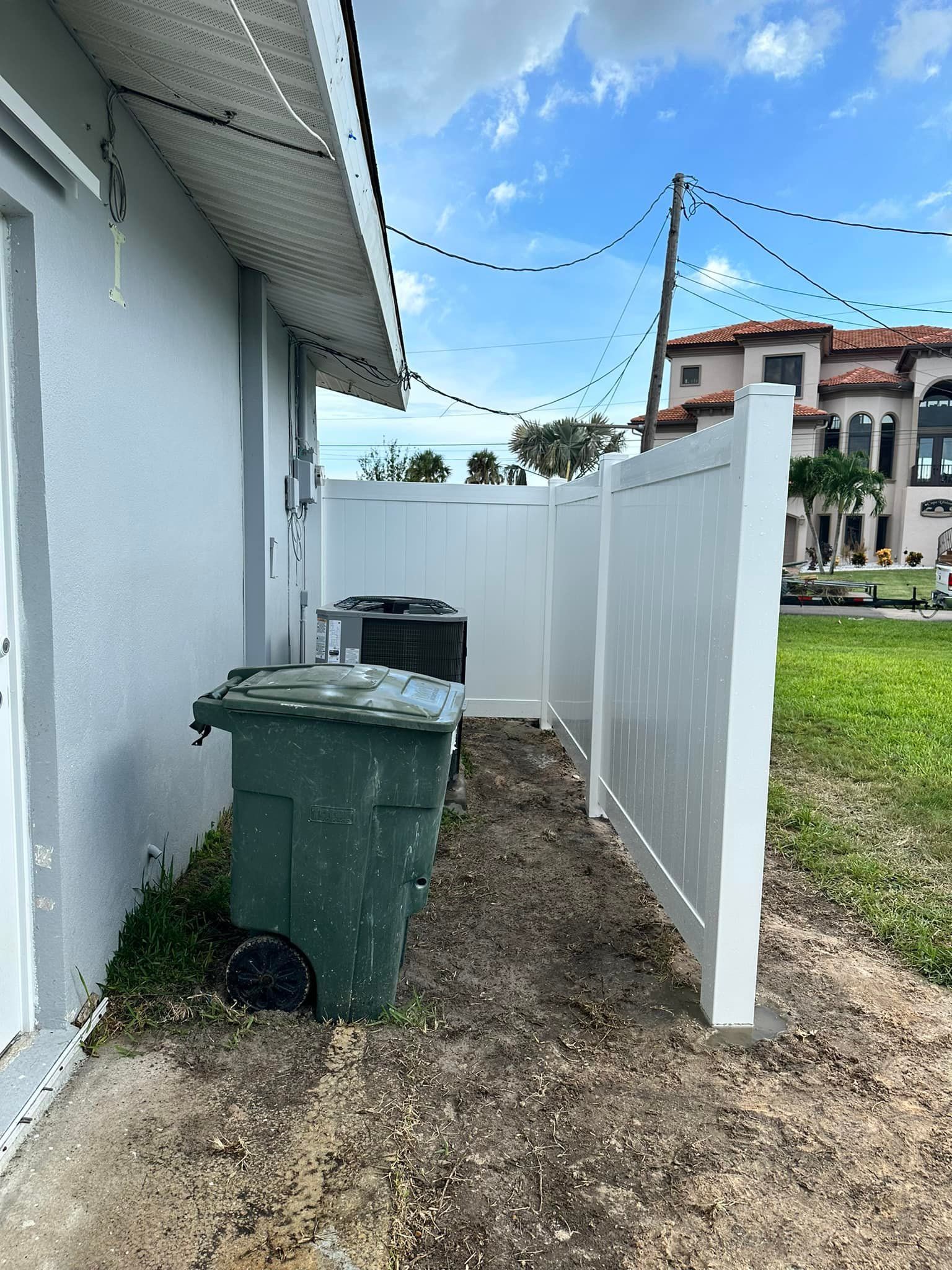 Green trash can, AC unit, and white fence along the side of a building. Grassy area under a blue sky.