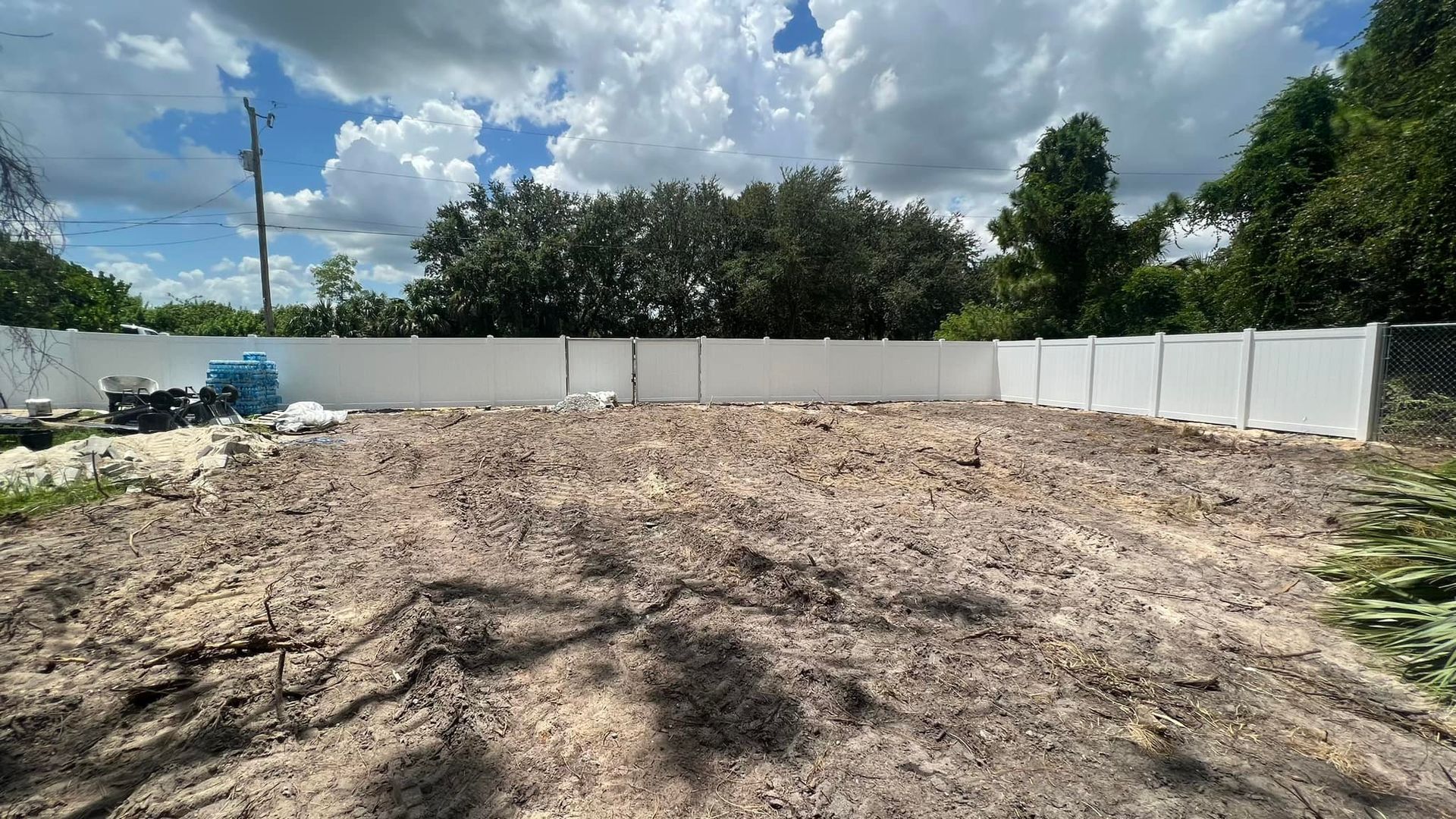 A dirt lot enclosed by a white fence under a blue sky with some trees.