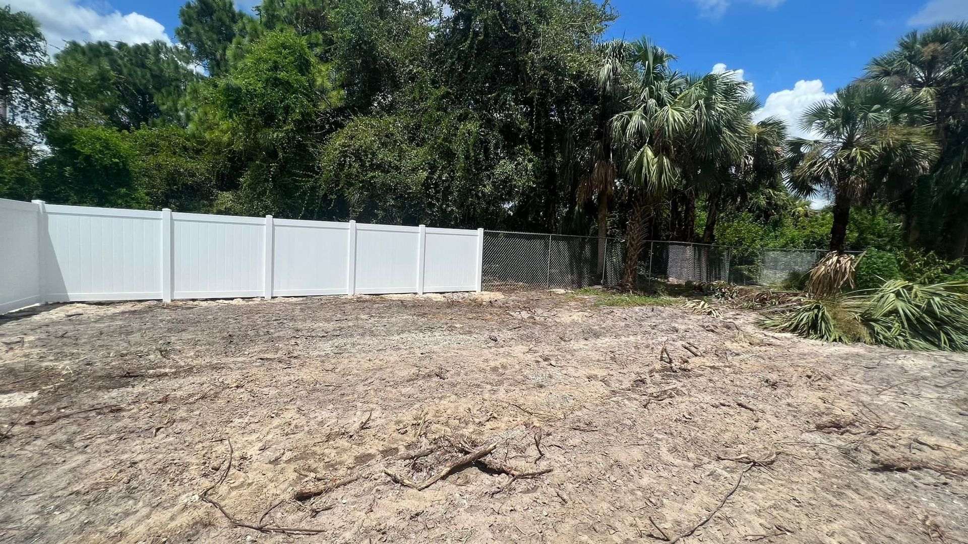 Cleared dirt lot with white fence and green trees in the background.