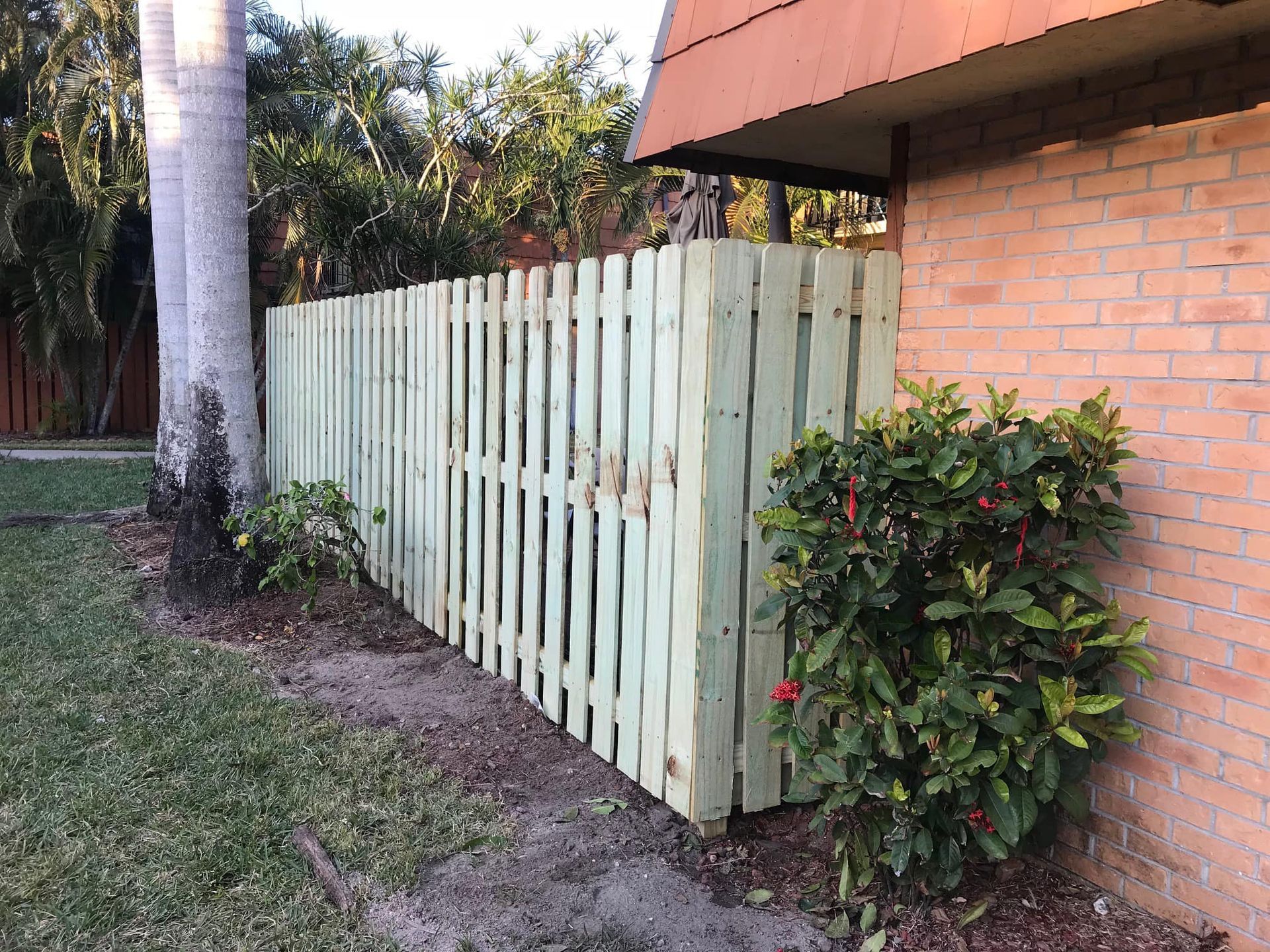 Green picket fence beside a brick wall and a palm tree, with shrubs and grass.