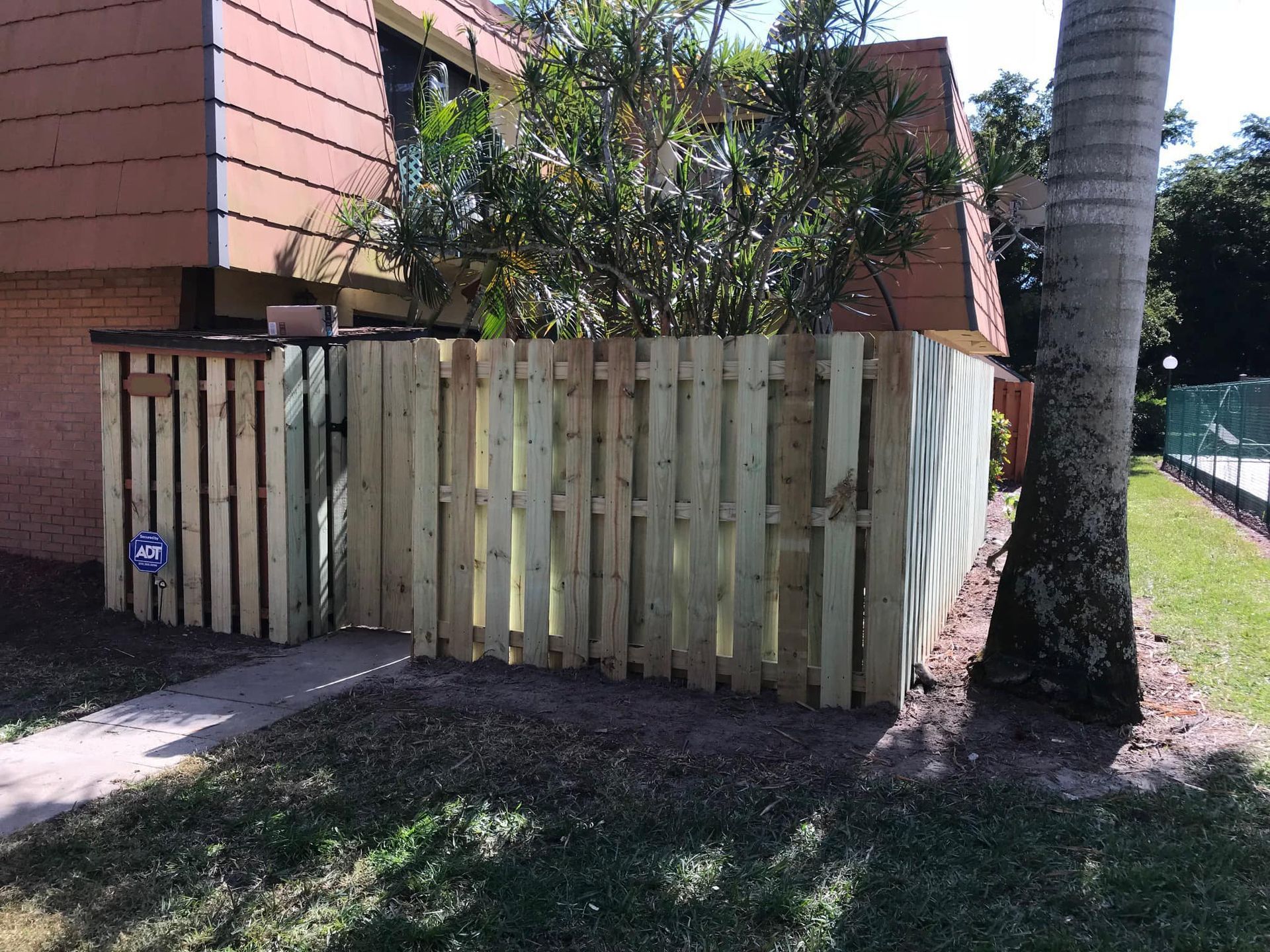 Wooden fence encloses a small area next to a building and tree. Green grass and pathway in front.