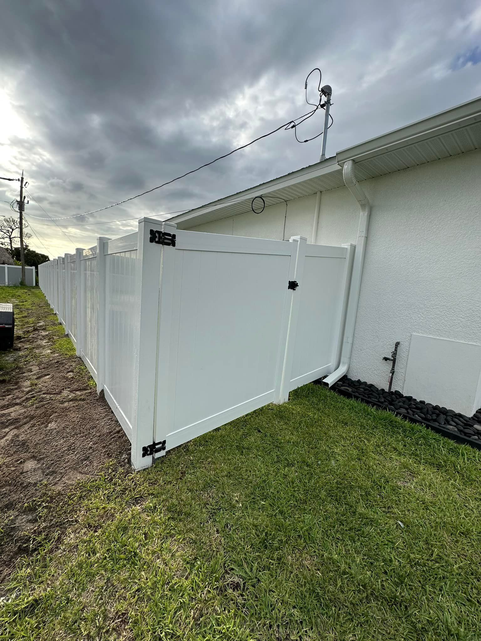 White vinyl fence with gate next to a white house, green grass. Overcast sky.