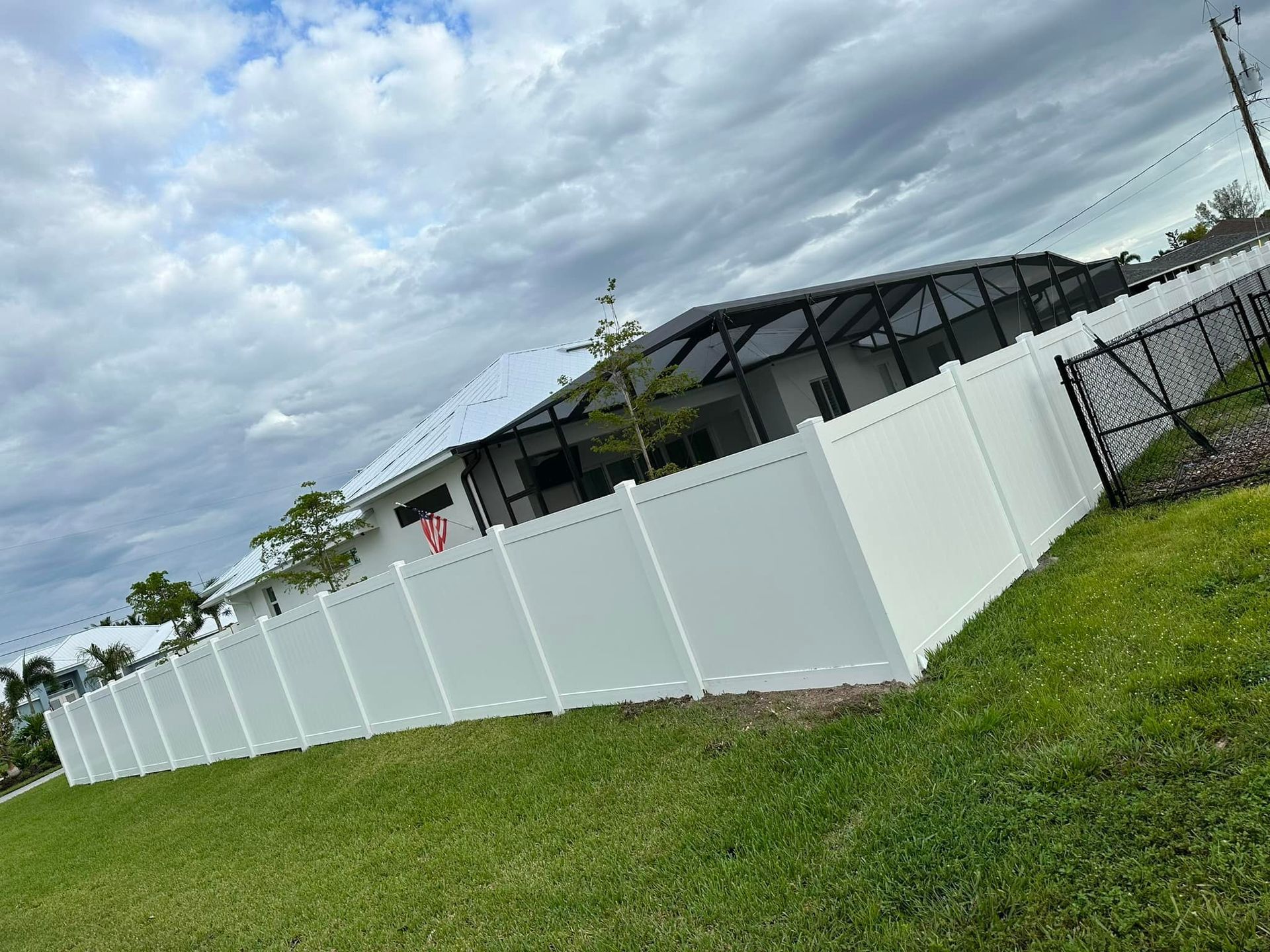 White vinyl fence surrounds a house with a dark screened-in porch, on a grassy lawn under a cloudy sky.