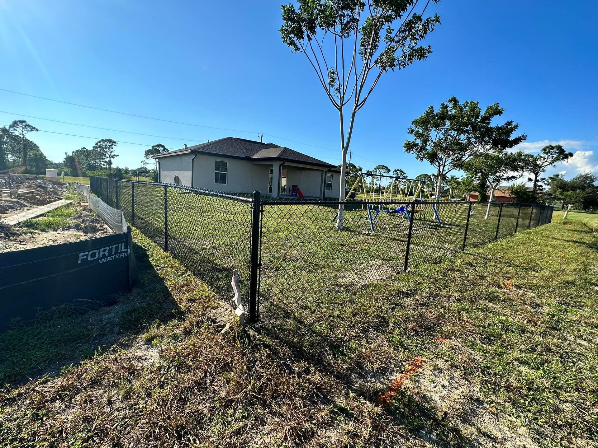 Black chain-link fence surrounds a grassy backyard with a beige house under a blue sky.