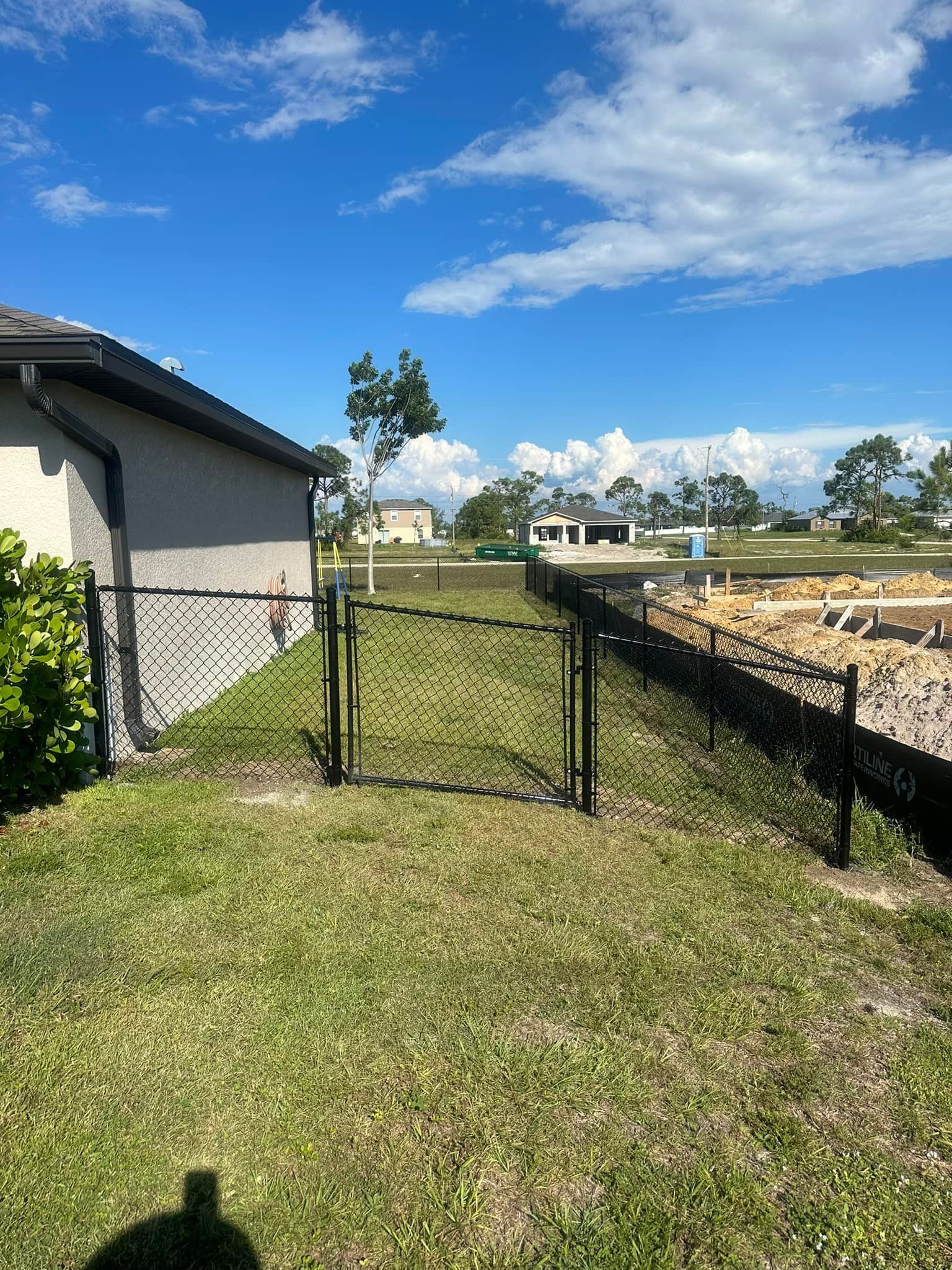 Chain-link fence with gate in grassy yard next to a house under a blue sky.