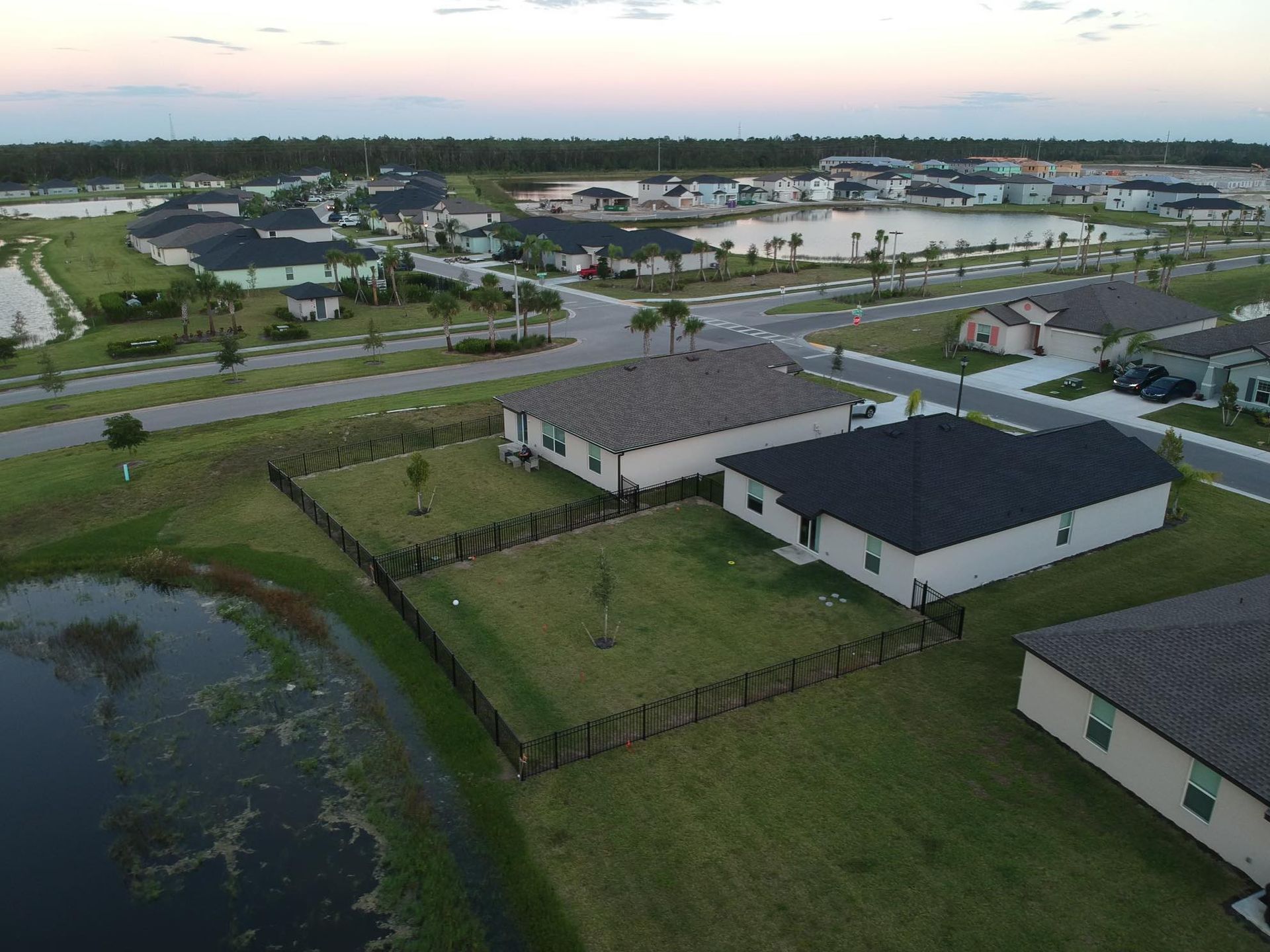Aerial view of suburban homes with dark roofs near a lake and roadways at dusk.