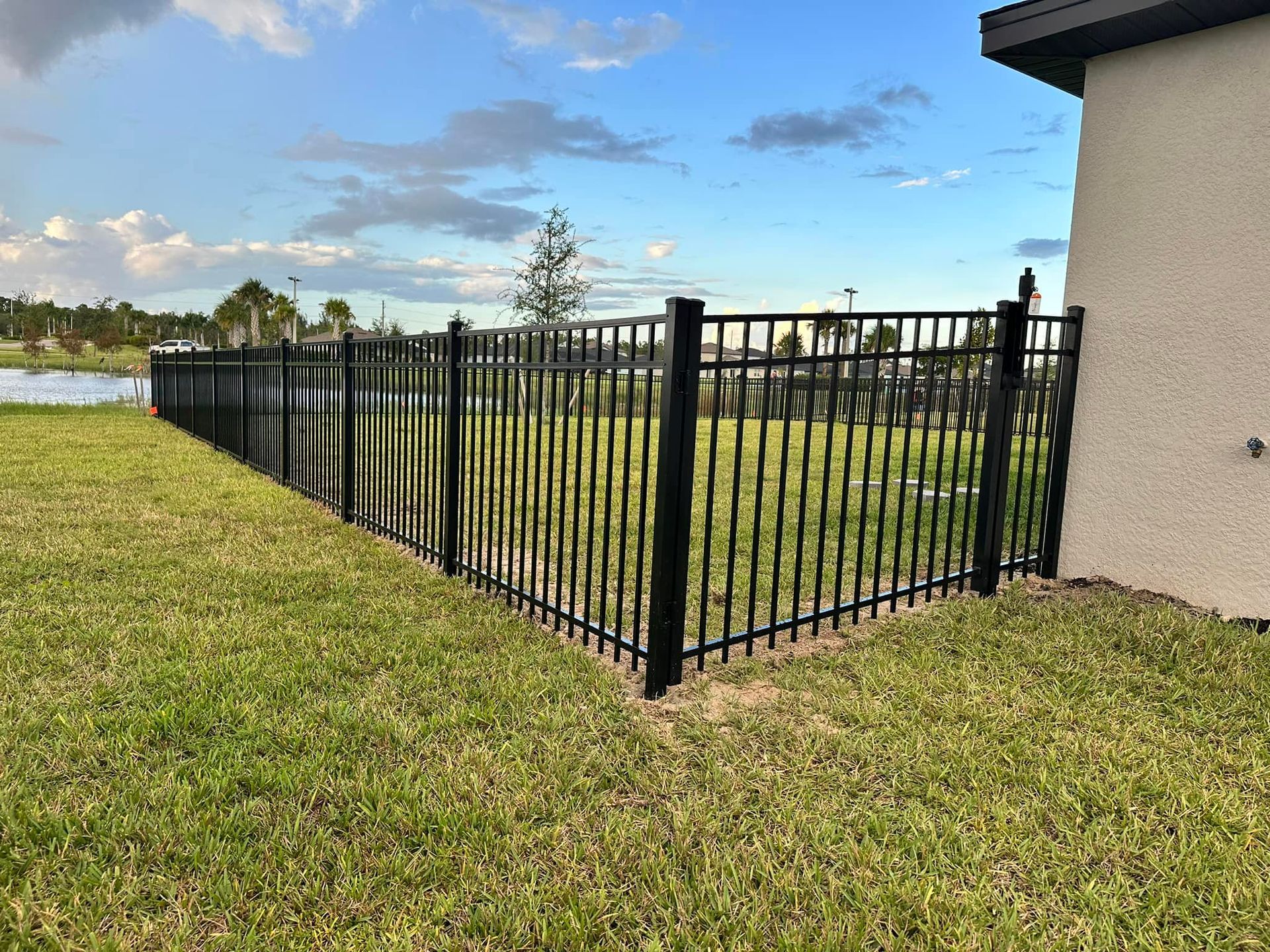 Black metal fence surrounds a grassy yard, next to a light-colored building, near water.