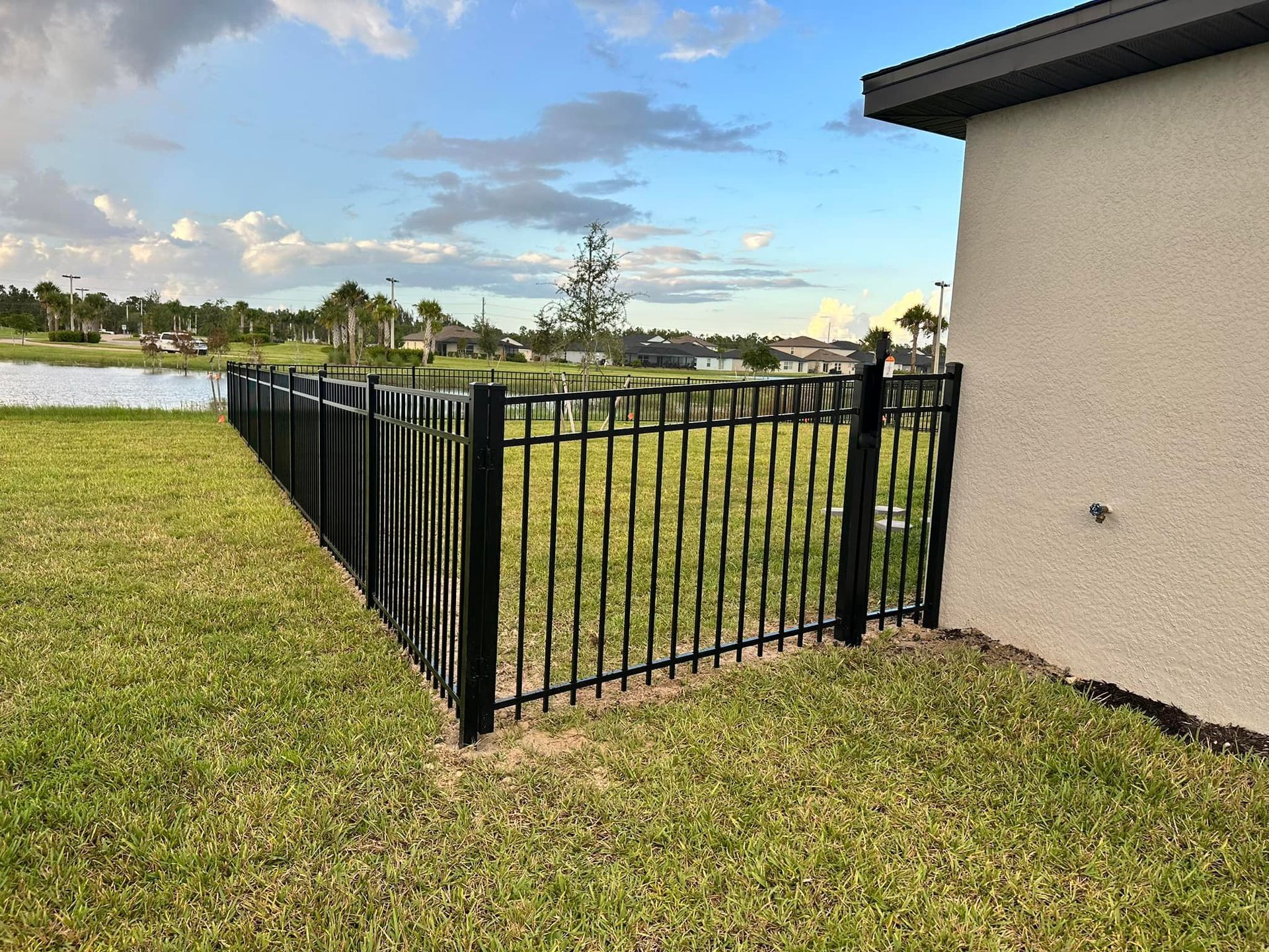 Black metal fence around a backyard, next to a tan house. Green grass, and water in the background.