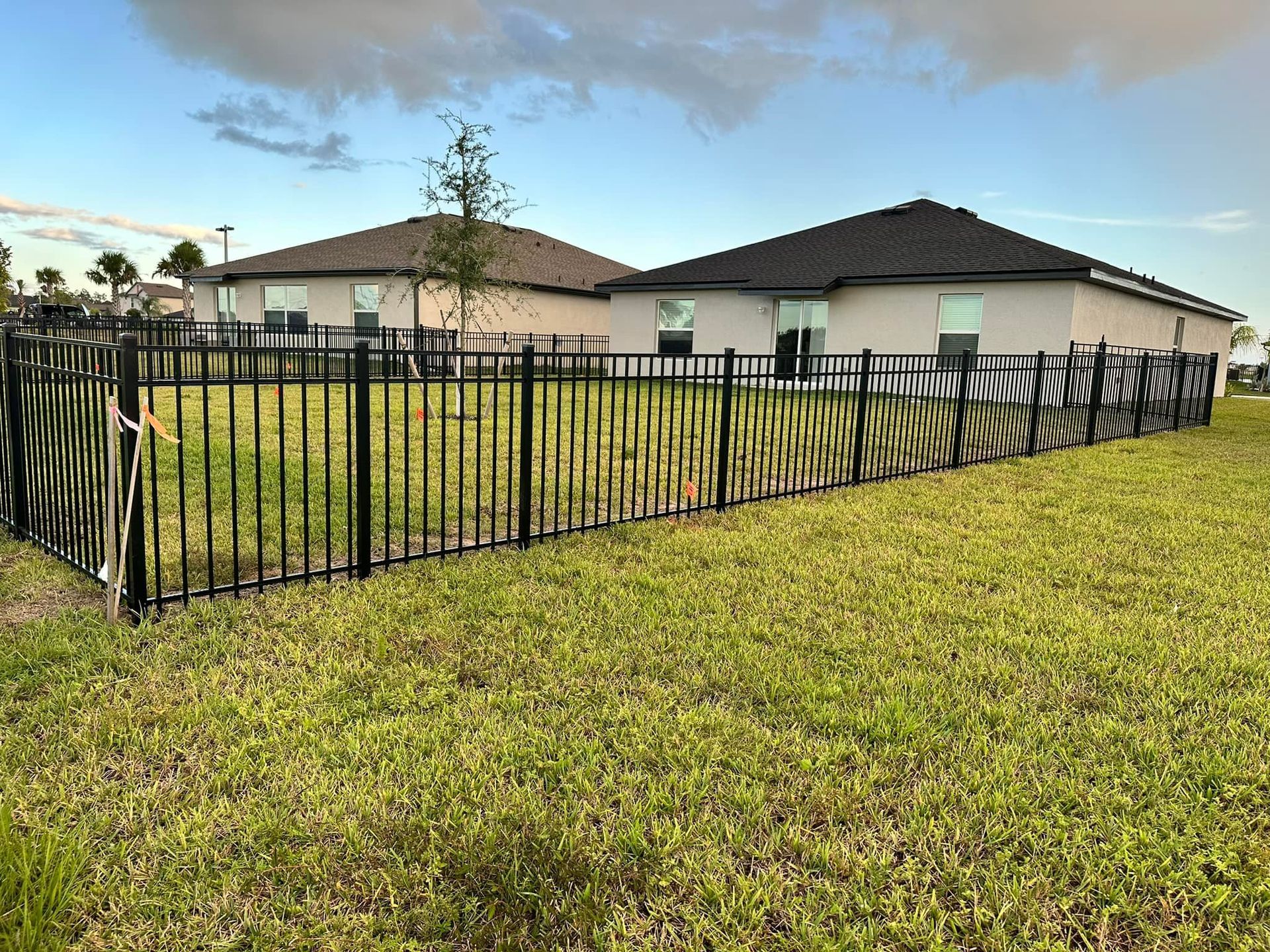 Black metal fence around a grassy yard, with beige houses in the background and cloudy sky.