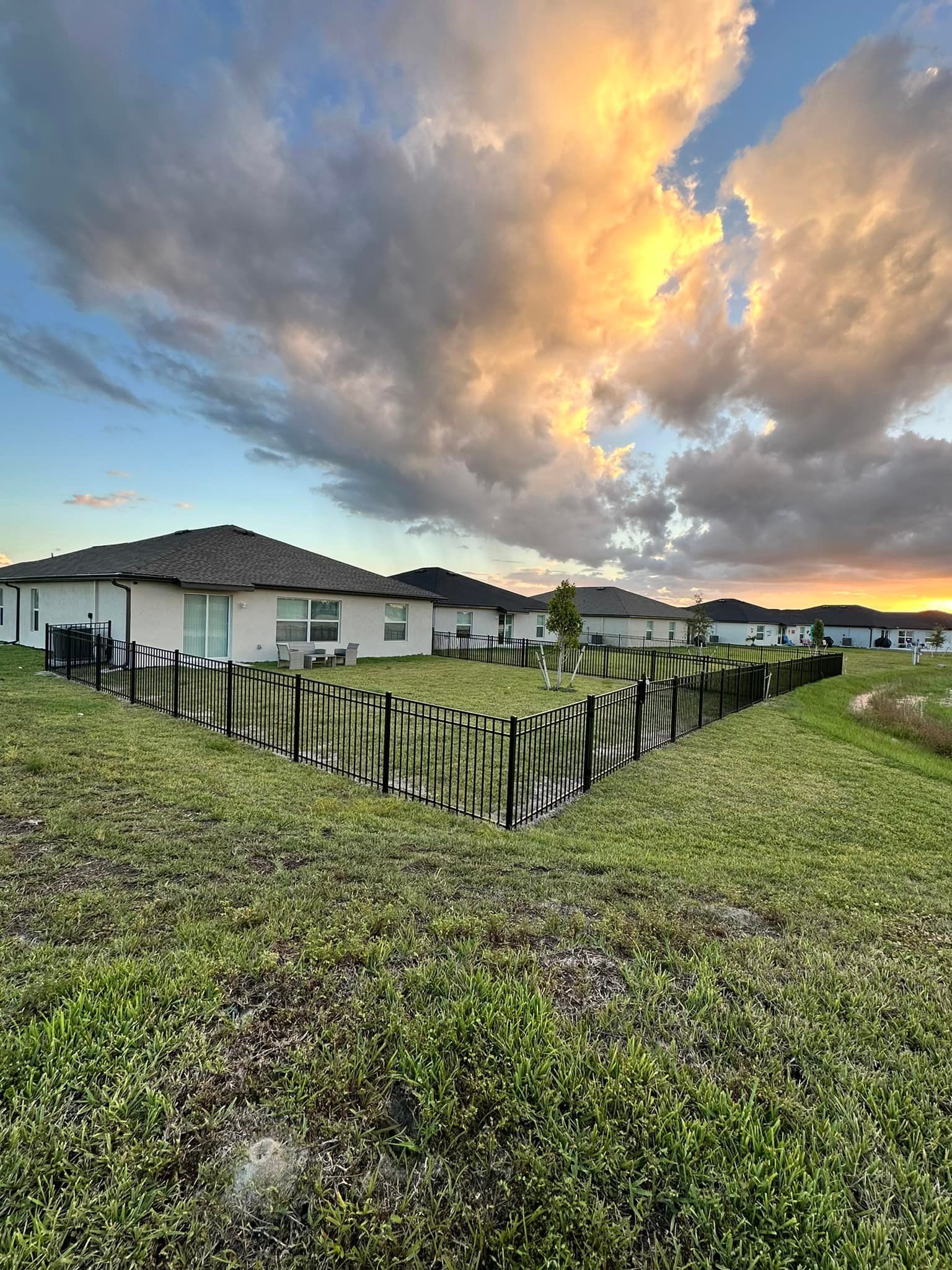 Backyard of a house with a fence, green grass, and a colorful sunset in the sky.