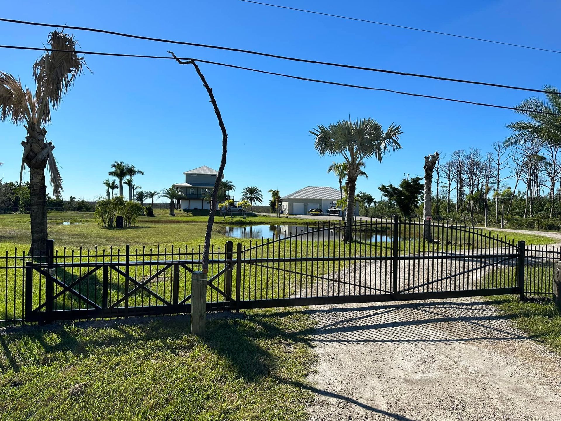 Black gated entrance to a home with a pond and palm trees under a blue sky.