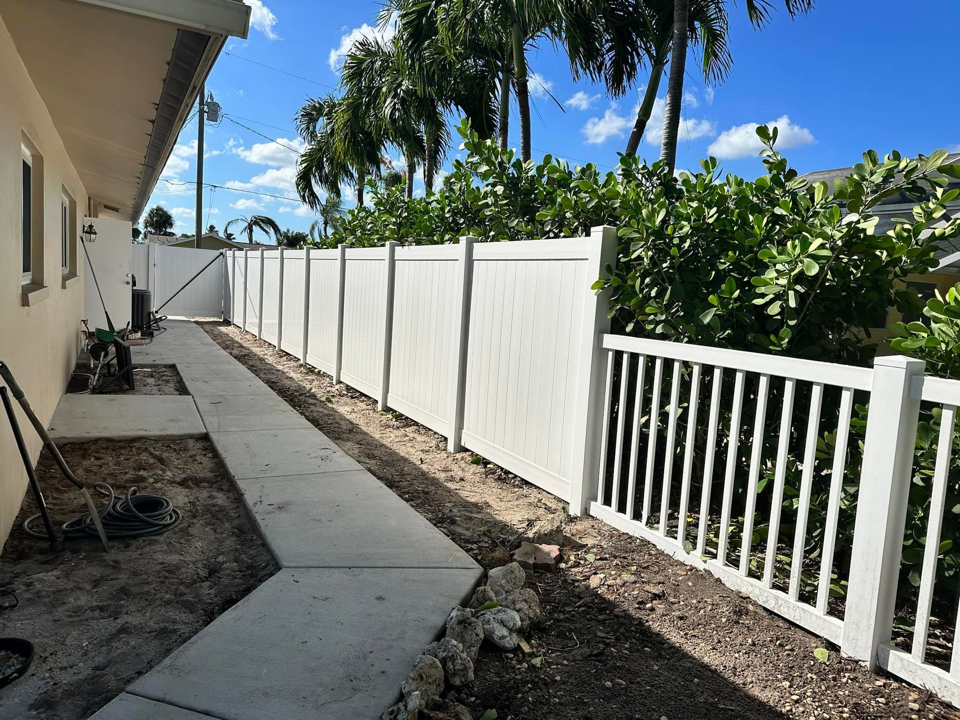 White vinyl fence alongside a concrete walkway and house, with lush green bushes and palm trees in the background under a blue sky.