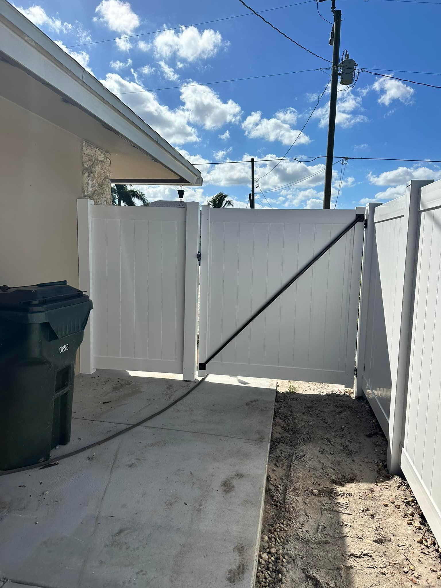 White vinyl fence with a gate, next to a house. A trash can sits nearby. Sunny day.