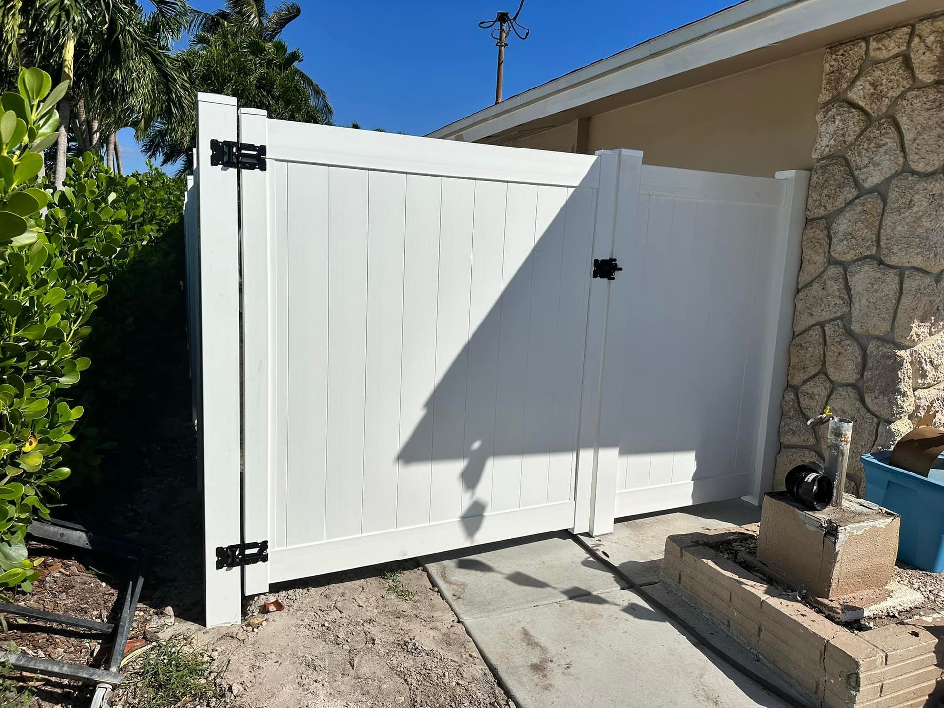 White vinyl gate with black hinges and latch, set in a sandy area beside a stone wall.