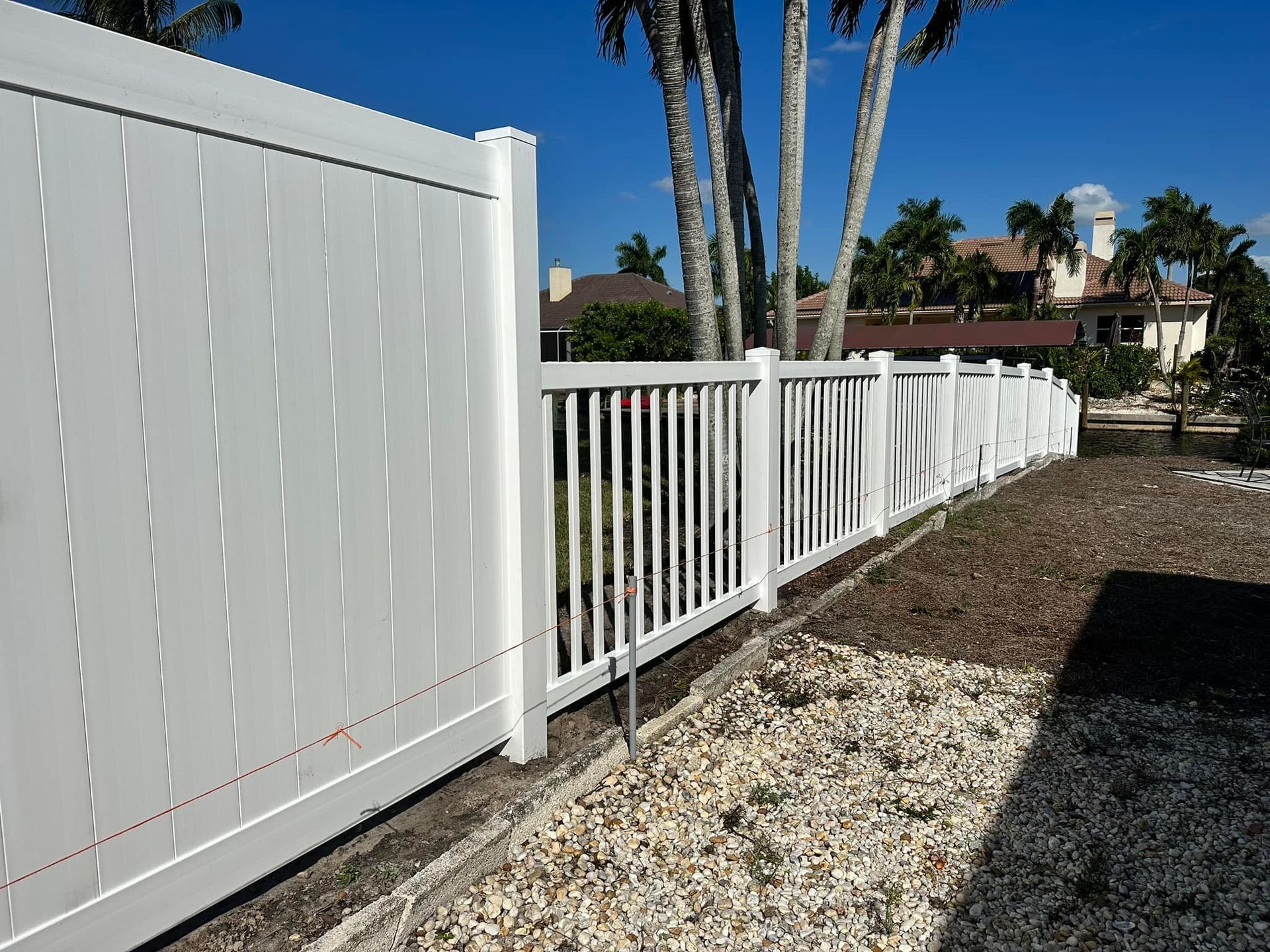 White vinyl fence in a sunny yard, with a partially visible house and palm trees in the background.