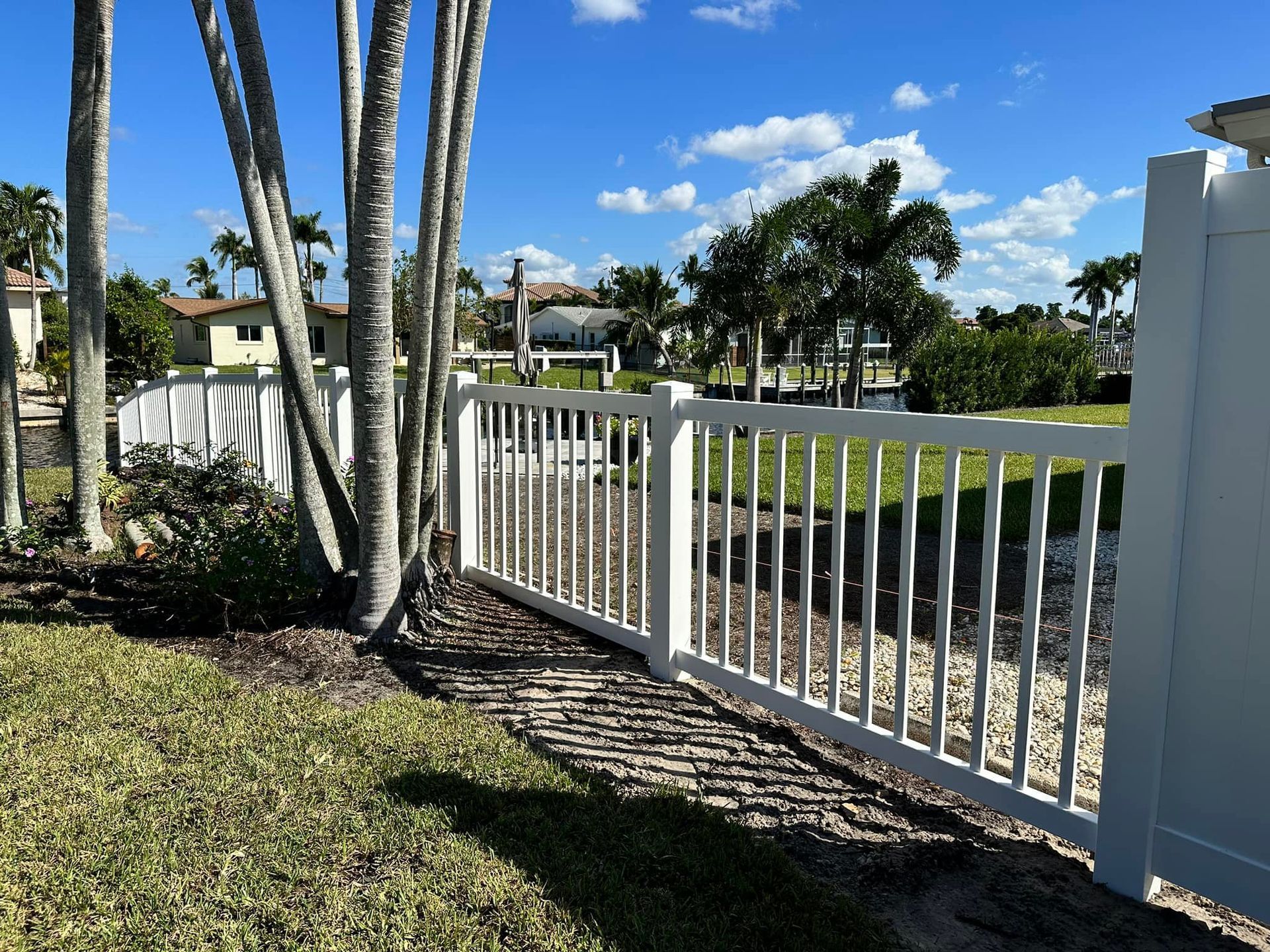 White fence bordering green grass and trees, with houses and blue sky visible in the background.