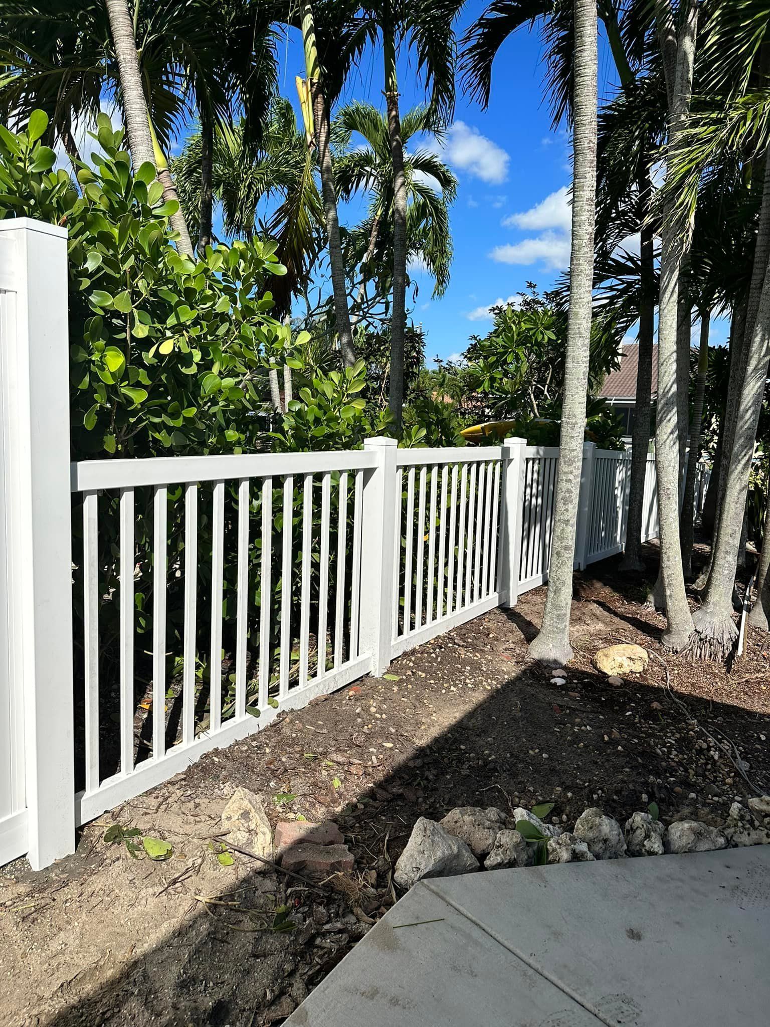 White picket fence bordering a yard with trees and a blue sky.