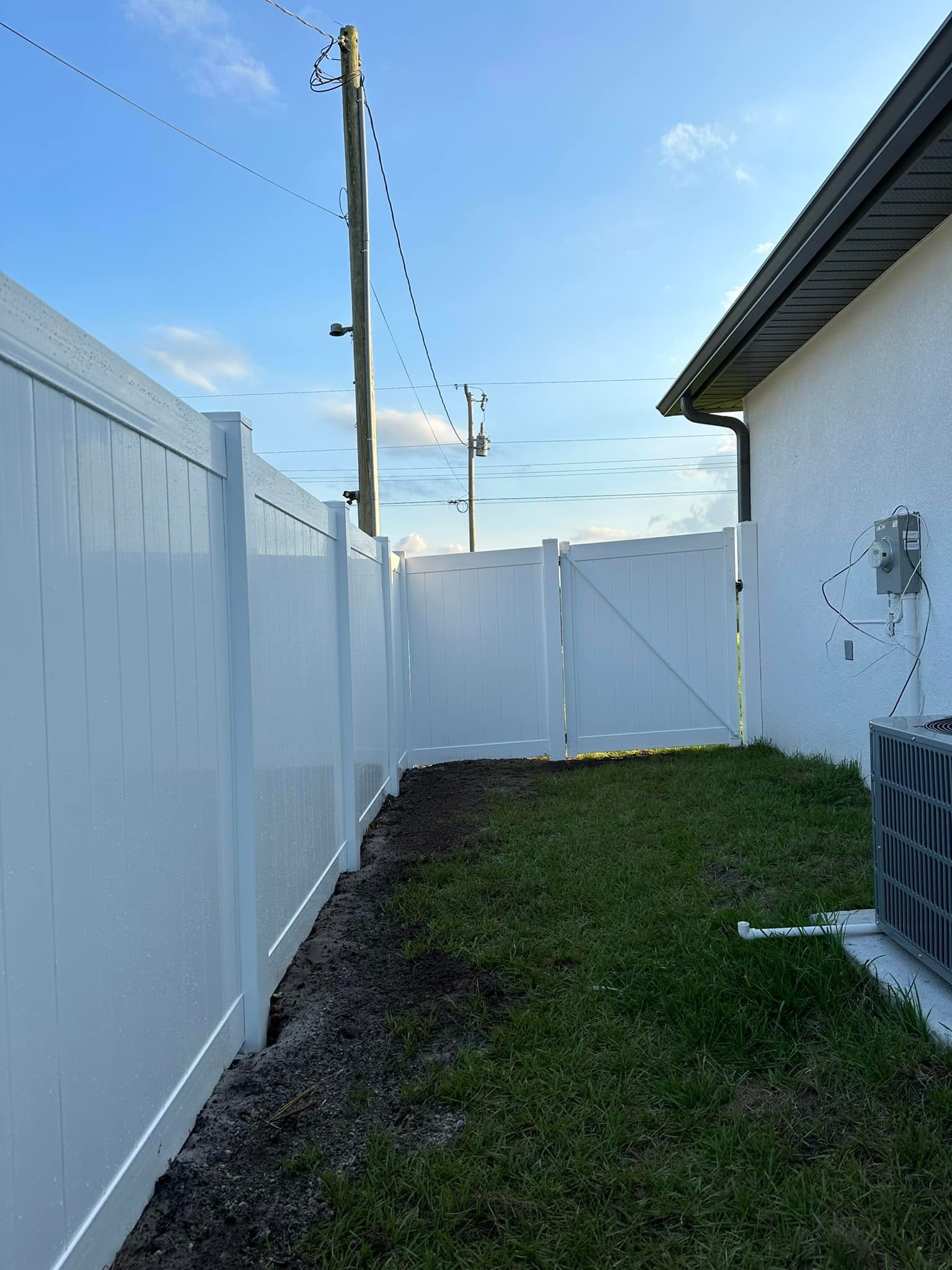 White fence with gate and grass area between house and fence, under a clear sky.