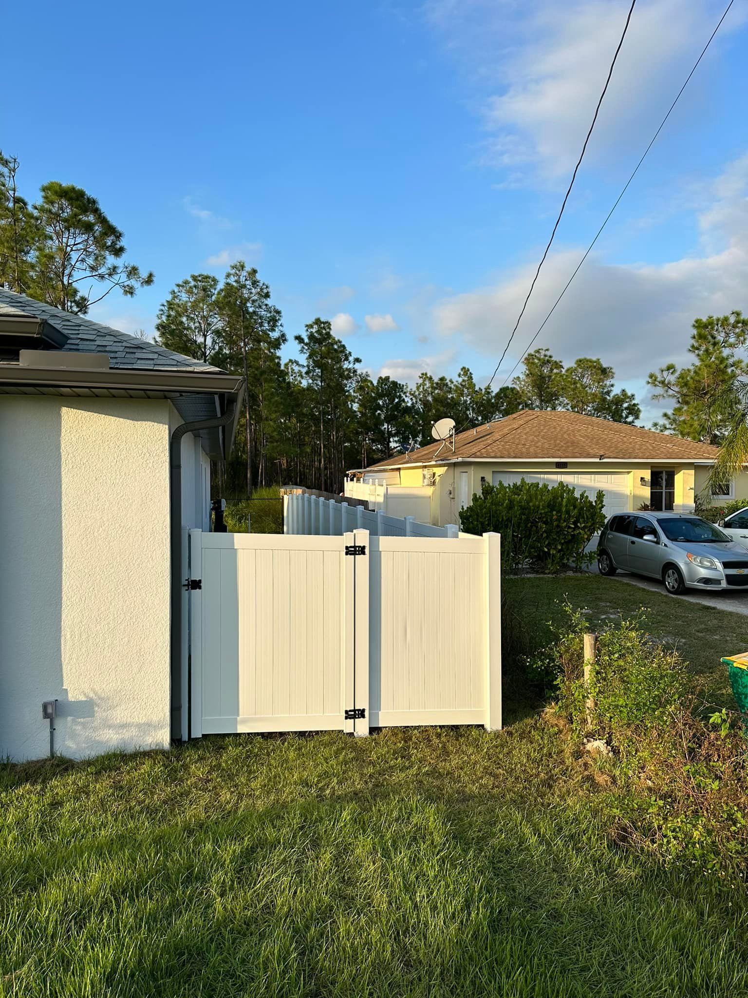 White fence gate next to a stucco building with a residential home in the background. Green grass, blue sky.