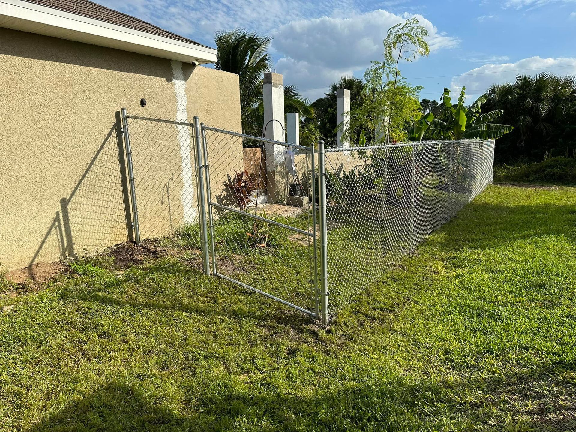 Chain-link fence next to a stucco house and green lawn, under a partly cloudy sky.