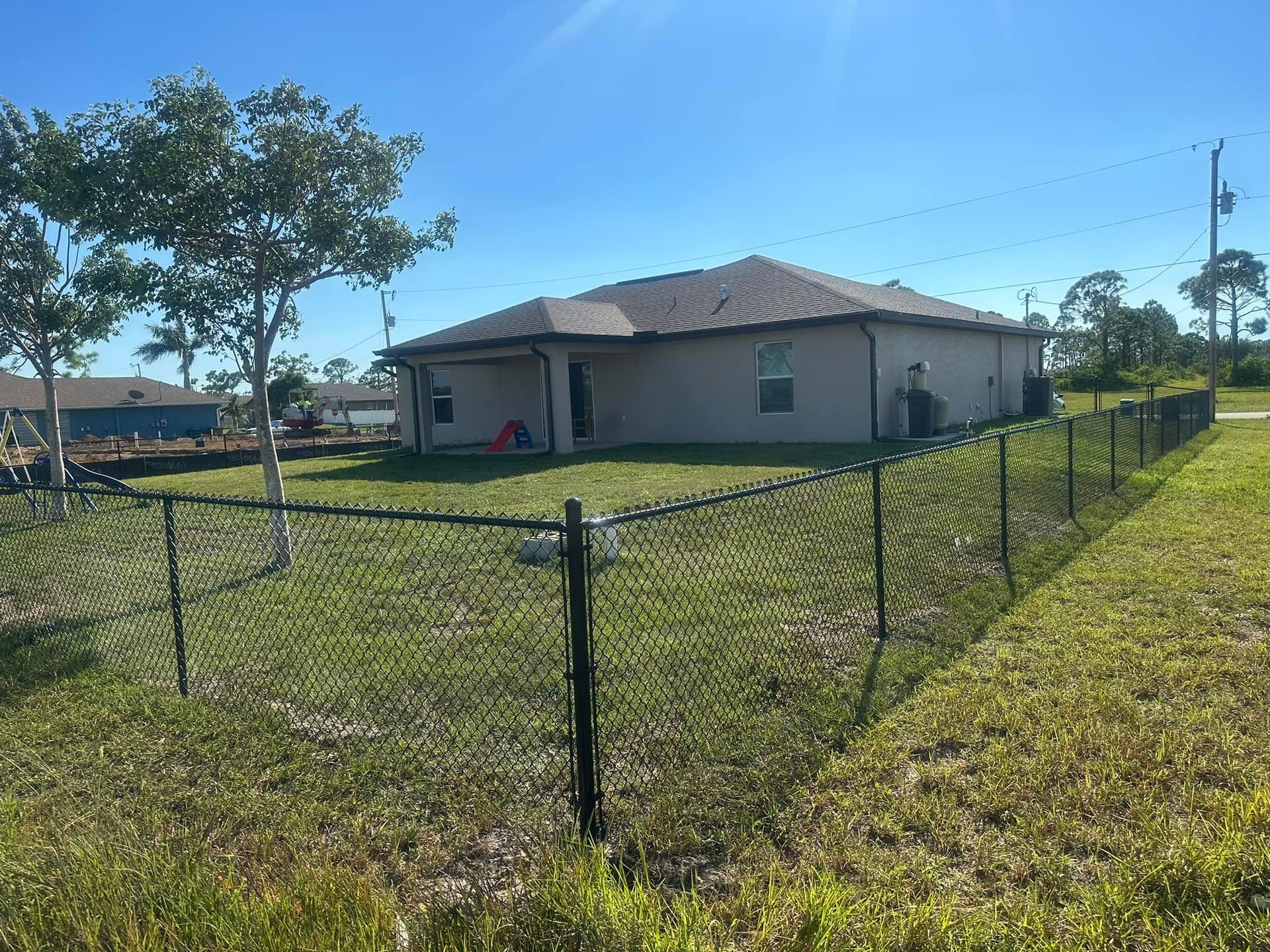 Black chain-link fence surrounds a light brown house with a dark roof, against a bright blue sky.