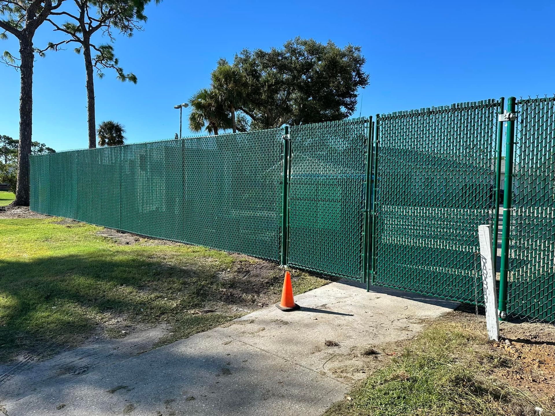 Green privacy fence with a gate on a sunny day. Orange traffic cone on the walkway.