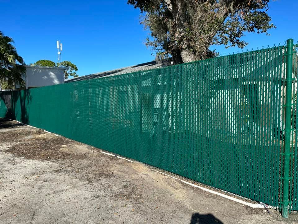 Green chain-link fence with privacy slats, surrounding a building on a sunny day.