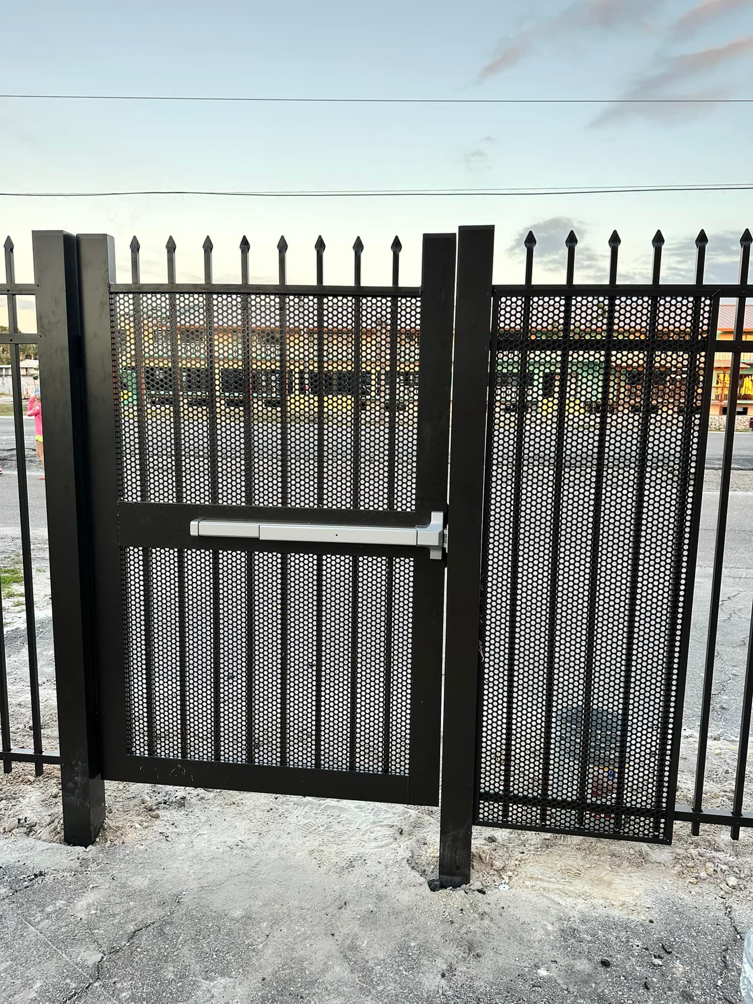 Black metal gate with perforated panels, set in a fence. Gray concrete ground.